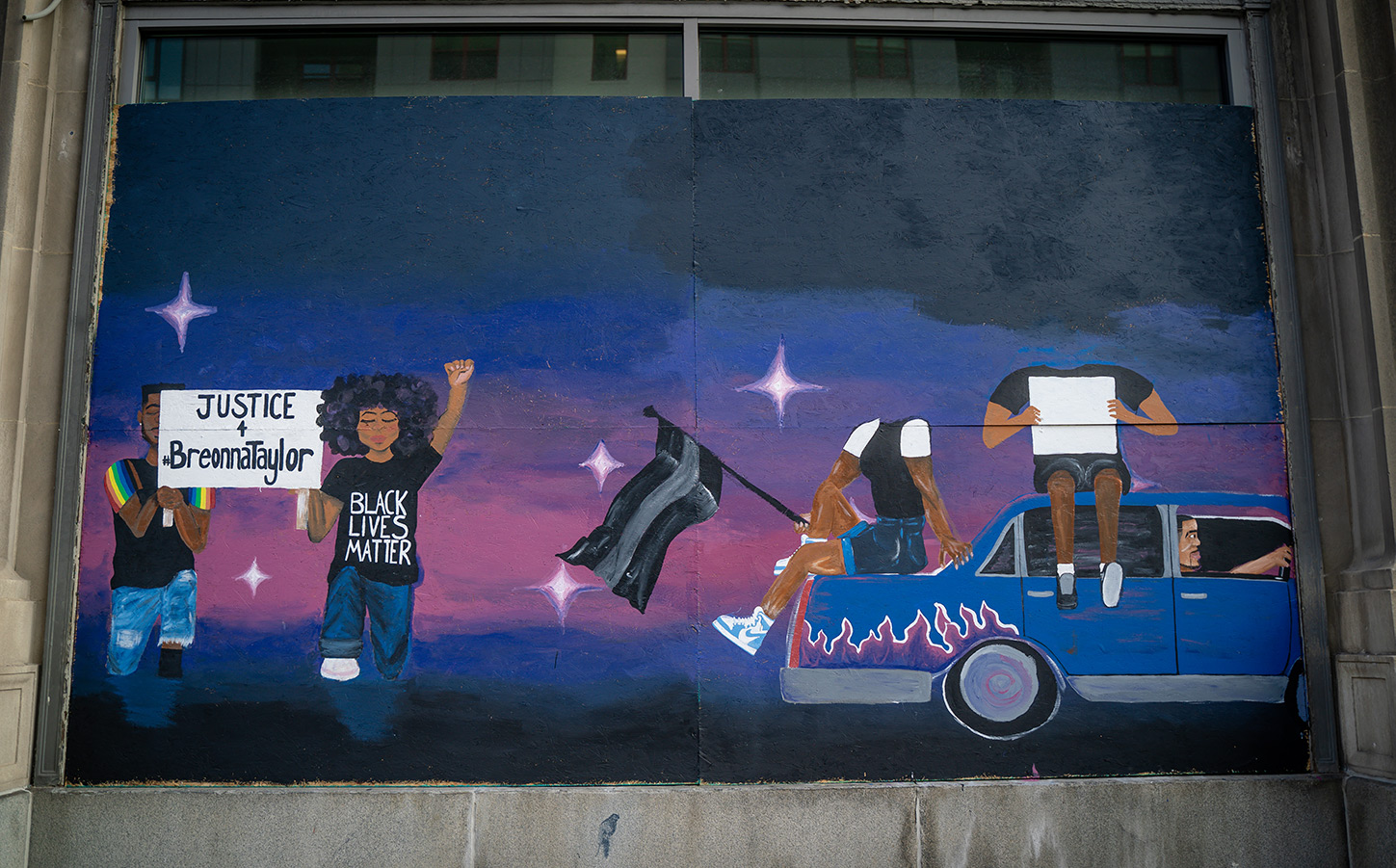 A color photo that is taken front-on. The picture is of street art painted on plywood covering windows on a building downtown Columbus. The background of the art is a top-to-bottom gradient: black, blue, pink, purple, blue, black. The bottom right corner shows two figures holding a sign that reads “Justice for Breonna Taylor.” The bottom right corner shows a blue car with pink and purple flames. Two headless figures sit on the car, third figure is driving the car in the rightward direction. 