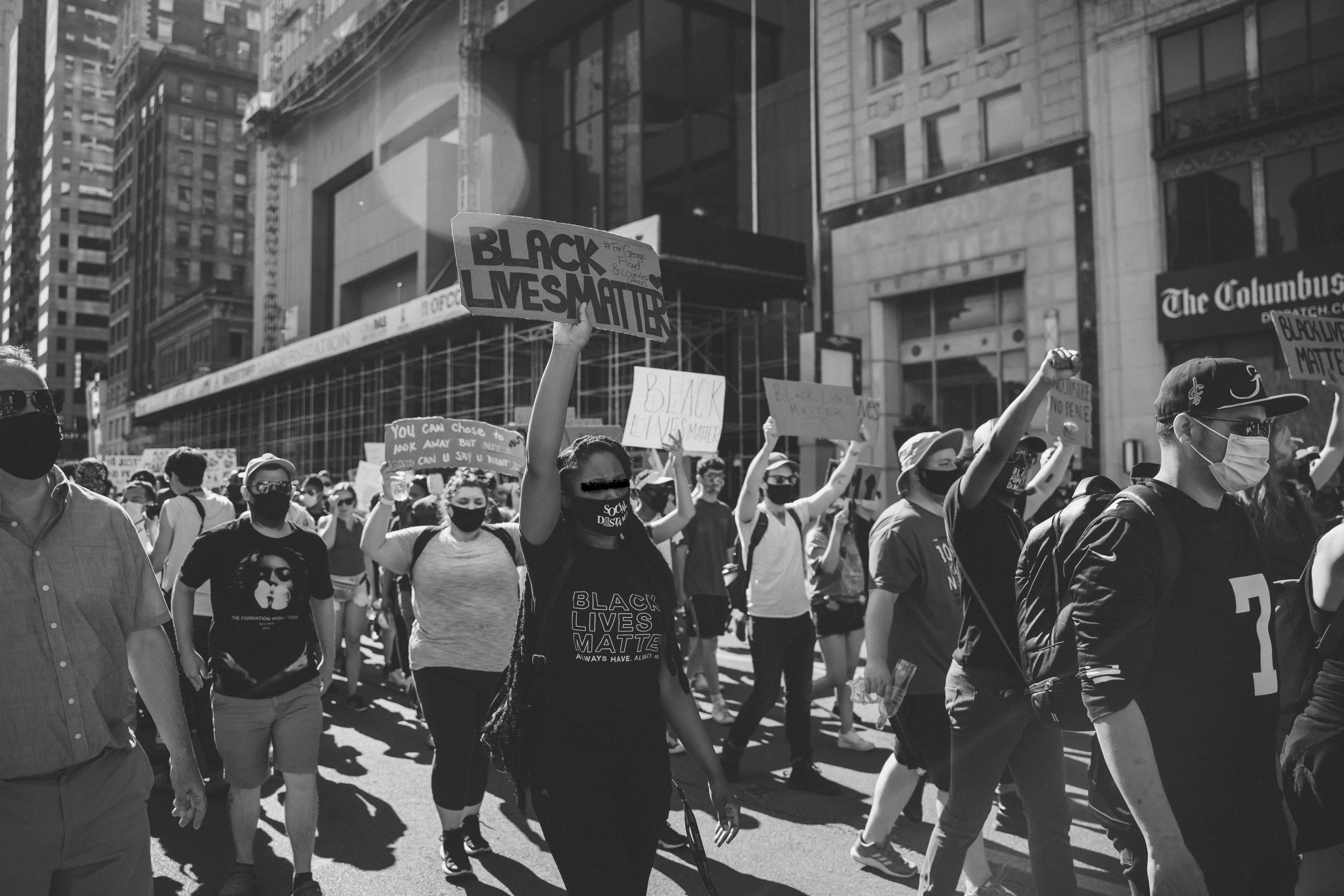 A black and white photograph with the bottom half of the frame fill of protestors demonstrating by walking from right to left, while raising their closed fists and holding signs. The focus predominantly lands on one protestor in the middle holding a sign and wearing a shirt that reads "Black Lives Matter."  