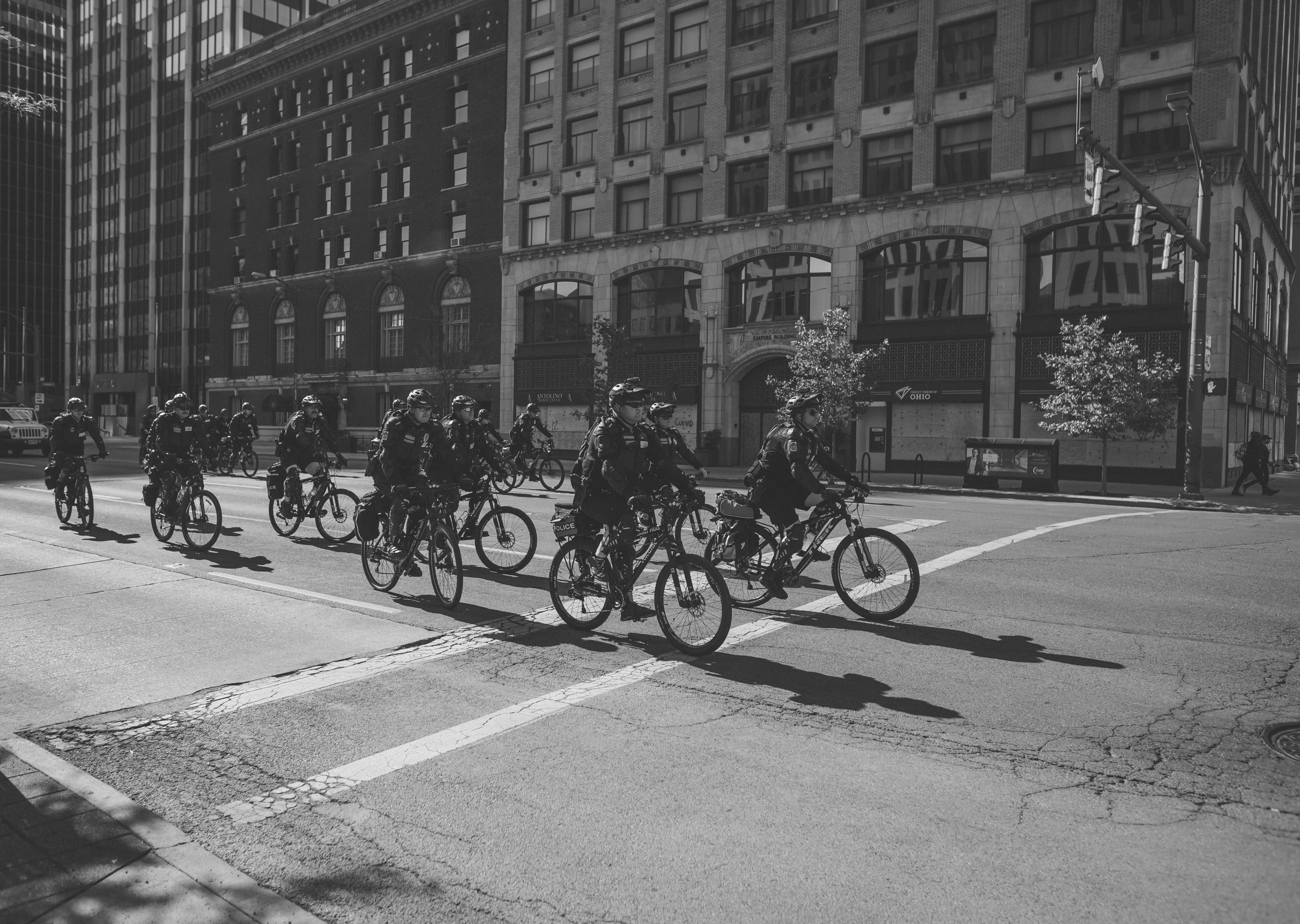 A black and white photograph. More than fifteen police officers on bicycles ride in the street from the right side of the frame and into the middle of the frame. They ride in three columns of about 5 officers. The background of the image shows buildings and the oncoming intersection.