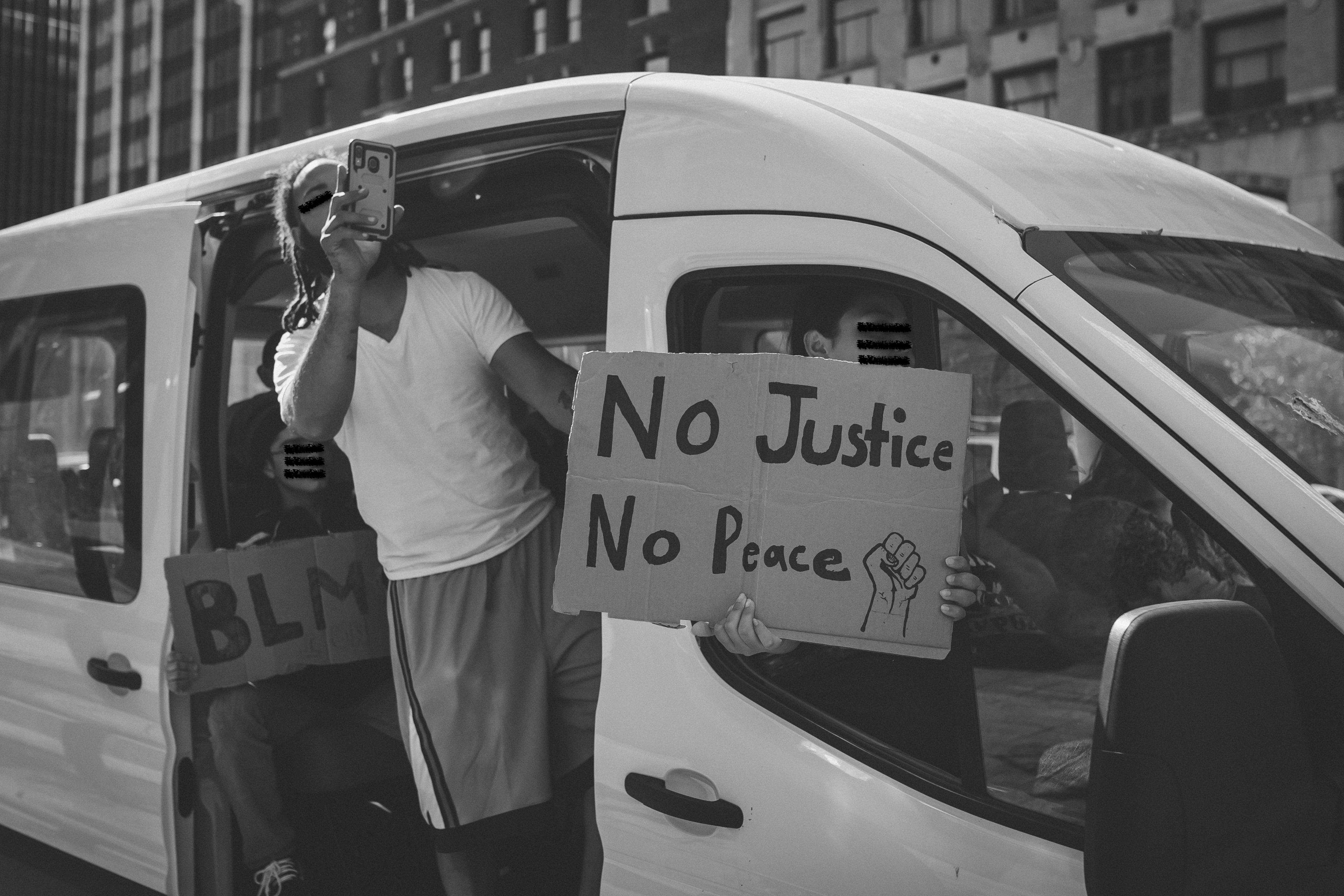 A black and white photo that fills the majority of the frame with protestors in a white van, shown from the passenger side. The van door is slid back and a protestor leans out of the door and holds up a phone to record. The shot-gun seat has a protestor that holds a sign out the window that covers their face and reads " No Justice, No Peace" with the image of a closed fist. In the back of the van, a third protestor sits and hold a sign that reads "BLM."  