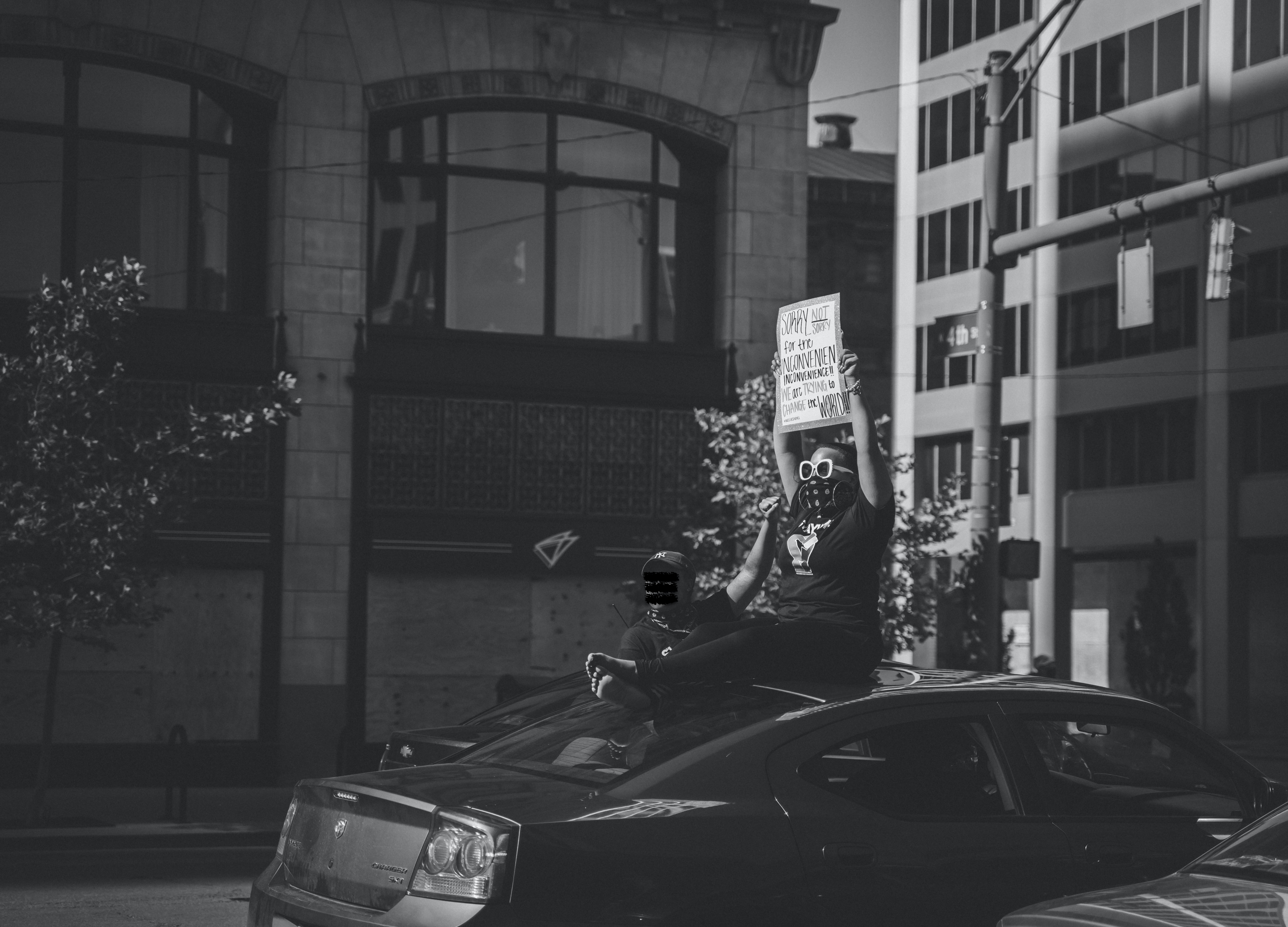 A black and white photograph. The foreground focuses on a BLM protester sitting on a car in the bottom right thirds of the image holding a sign that reads "Sorry, not sorry for the inconvenient inconvenience!! WE are TRYING to CHANGE the WORLD!" A second figure stands to the side of the car farthest from the viewer and raises a closed fist. The background shows buildings and stop lights.