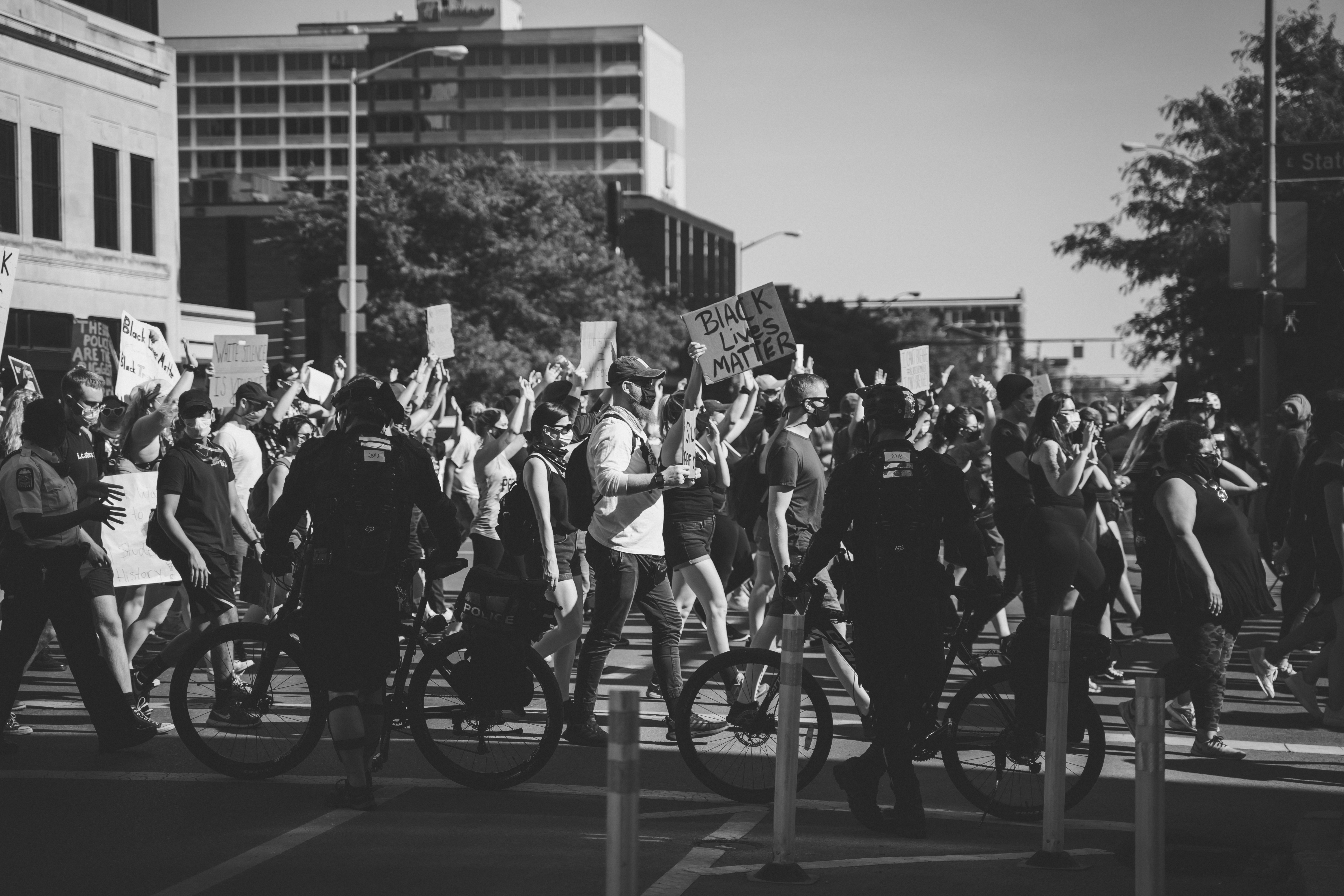 Black and white photograph shows Black Lives Matter protesters walking in one direction (from left to right) and consuming the bottom of half of the image, some of them raise signs. Between the viewer and the protestors are two police officers standing next to their bicycles with their backs turned to the viewer. The top half of the fame shows the sky, trees, and the tops of buildings.