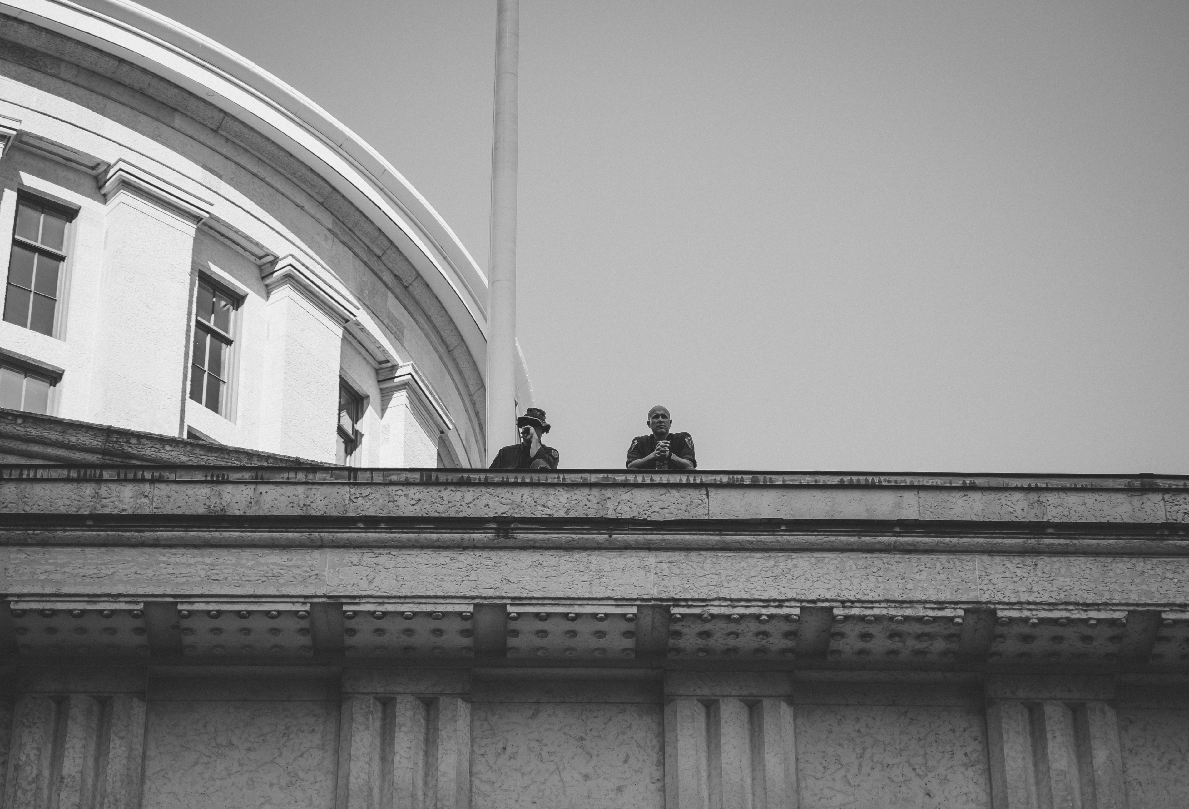 A black and white photo. In the middle of the image are two police offers overlooking BLM protests at the Ohio Statehouse. The viewer stands beneath them and sees the officers from the elbows up. The top right corner os the frame shows the dome of the Statehouse and the reminder of the top half shows and even gray-toned sky.