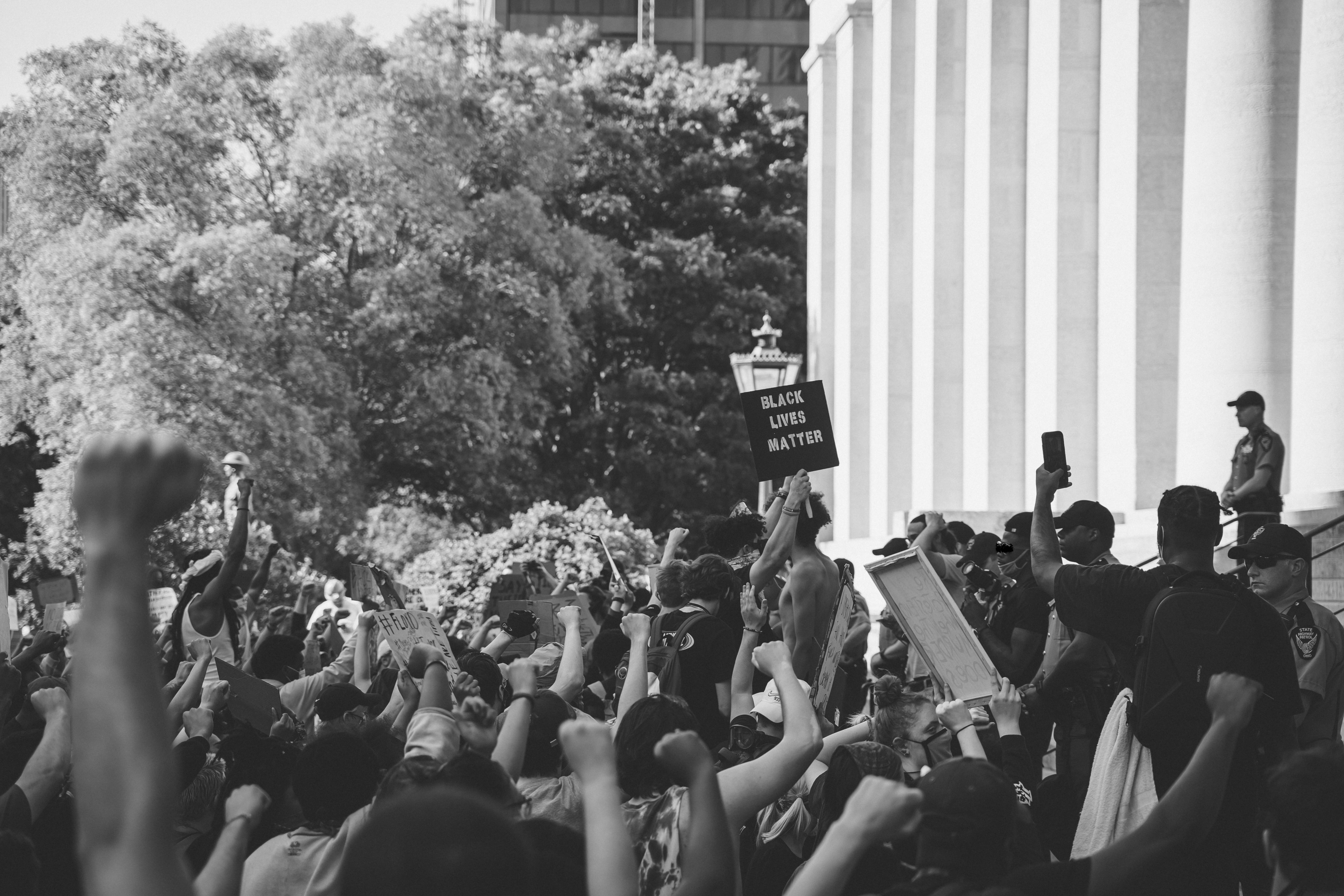 Black and white photo shows BLM protestors at Ohio State house. The viewer stands behind the protestors on their right-hand side. Majority of the protestors have bare arms and raised closed fists or signs and fill the bottom third of the image. There is one readable sign: "Black Lives Matter." The pillars of the state house fill the right half of the image above the protestors. The top left half of the image shows leafy trees.