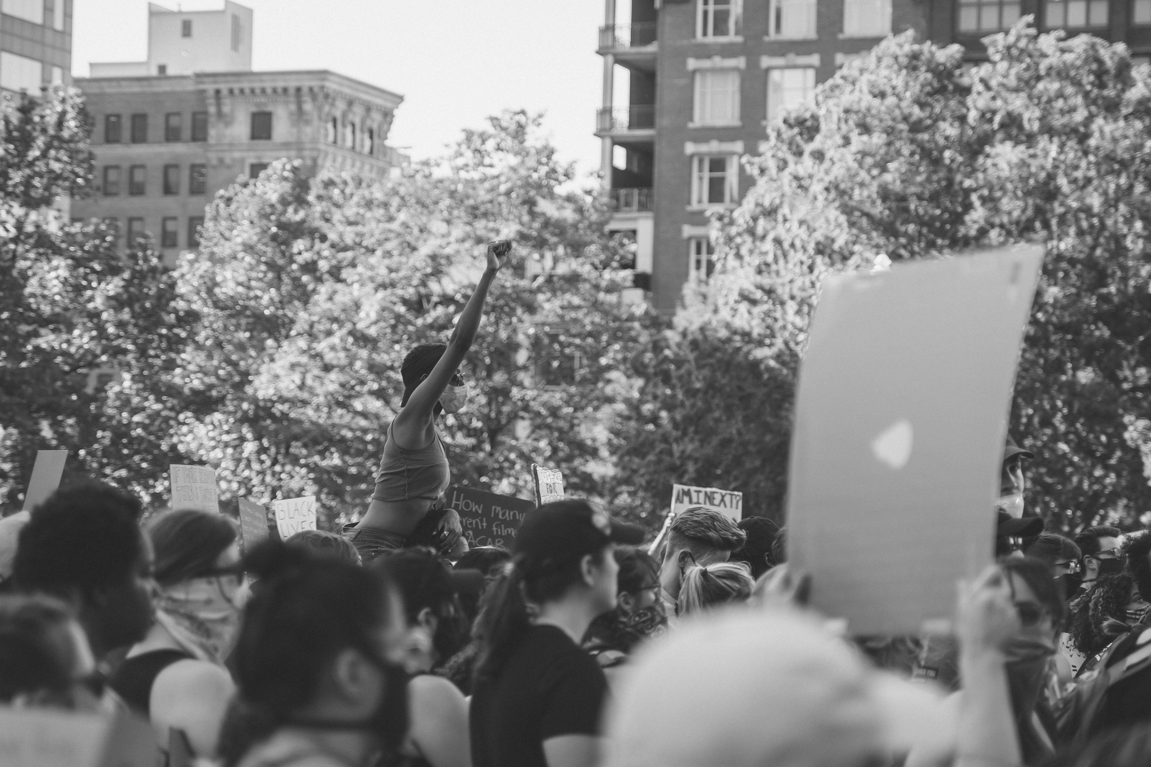 The middle ground of the black and white photo is in focus and shows a protestor during black lives matter demonstration sitting on someone's shoulders and is raising their closed right fist. The foreground shows blurred protestors facing the right side of the photo, with some holding signs. The background shows trees and buildings.