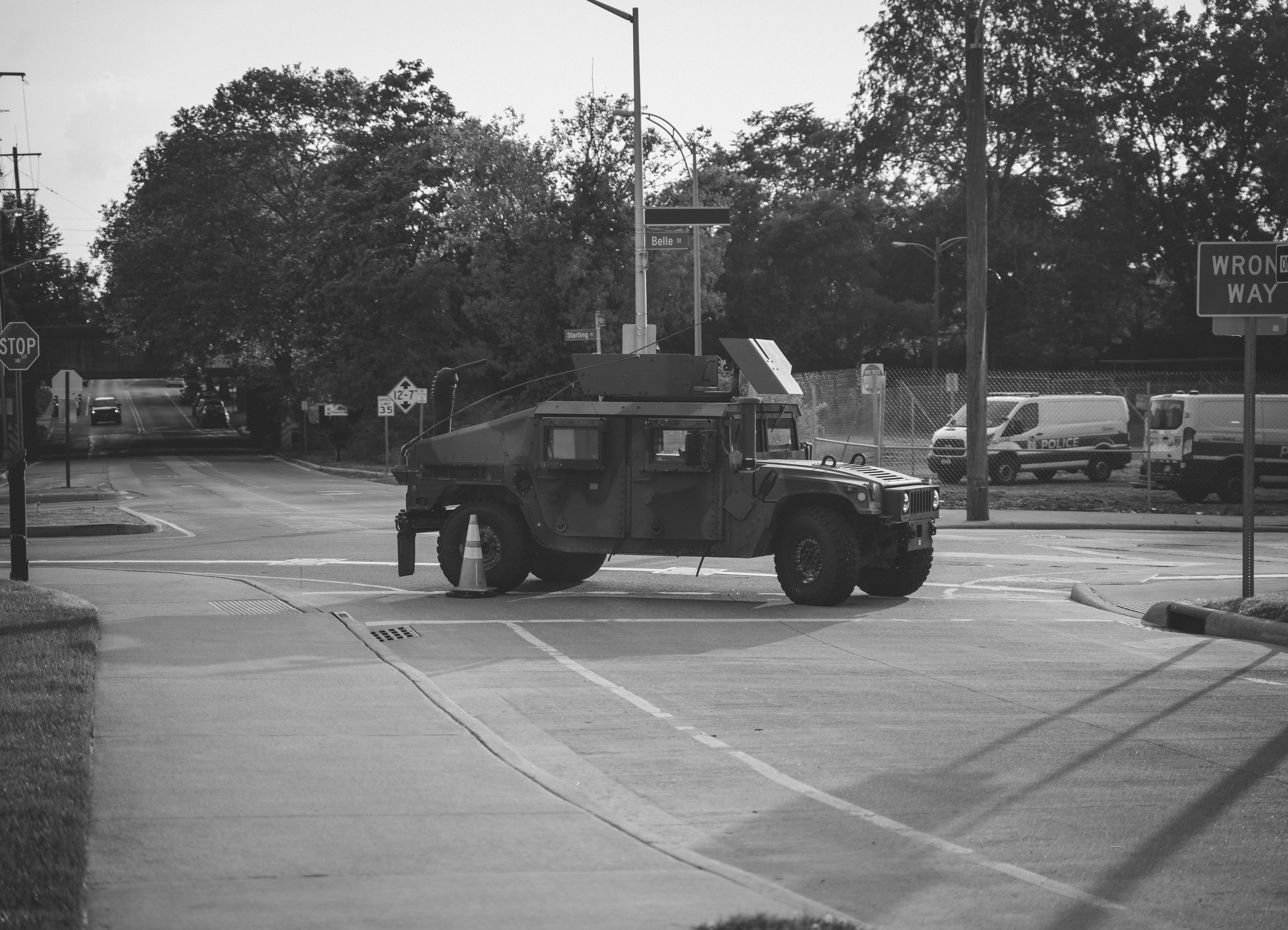 A black and white photograph. The center of the frame is a military vehicle in downtown Columbus during BLM protests and blocks off an intersection. The background features police vehicles, streetlights and four roads that meet at the intersection the military vehicle sits in. On the right edge of the image, a street sign that is cut off a bit reads "Wrong Way."