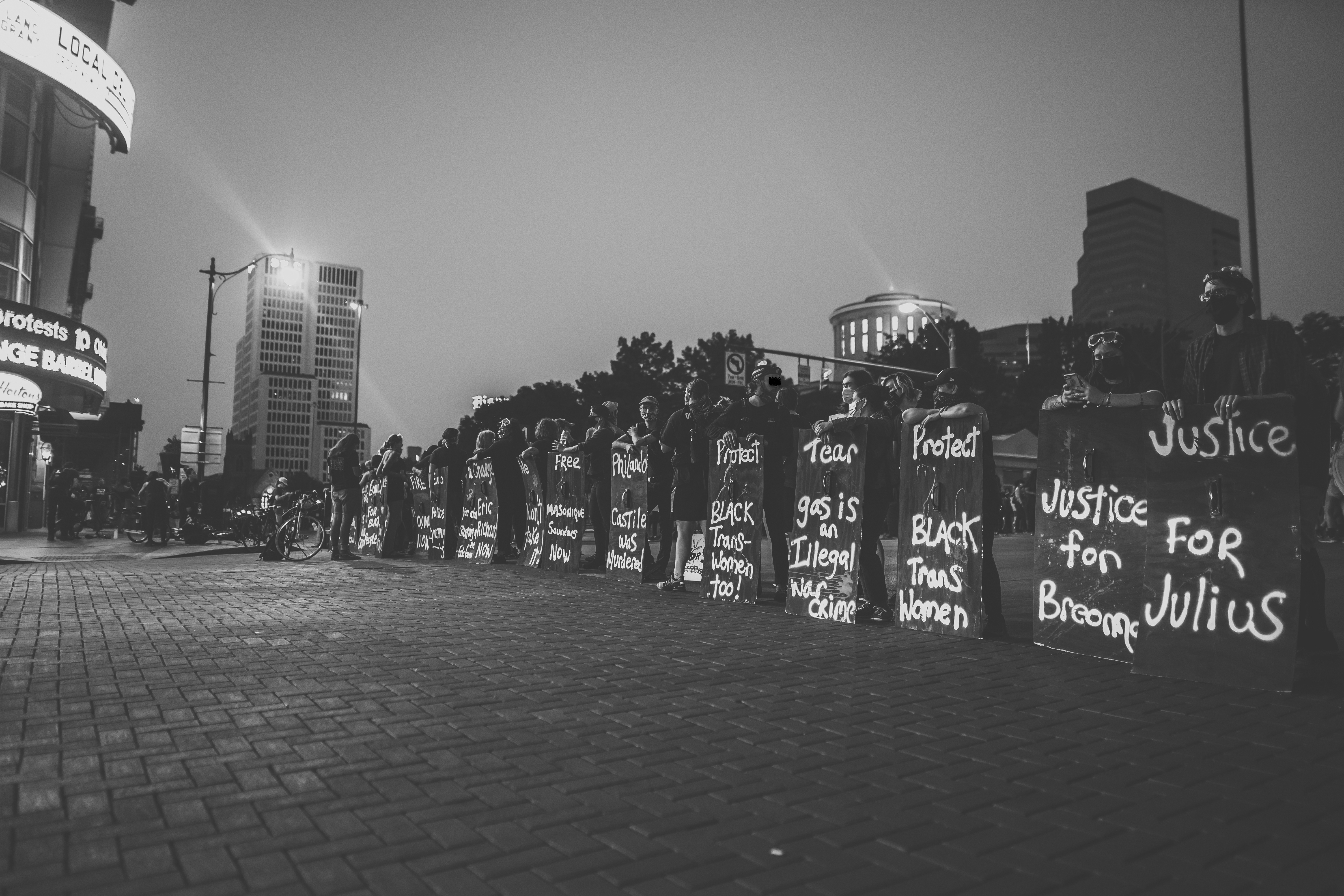 A black and white photograph that shows BLM protesters lined up from the bottom right corner of the image and receding into the background in the middle of the frame. The protestors stand behind signs that reach chest-height in downtown Columbus. Some read: "Justice for Julius," "Protect Black Trans Women," or "Tear gas is an Illegal war crime." 