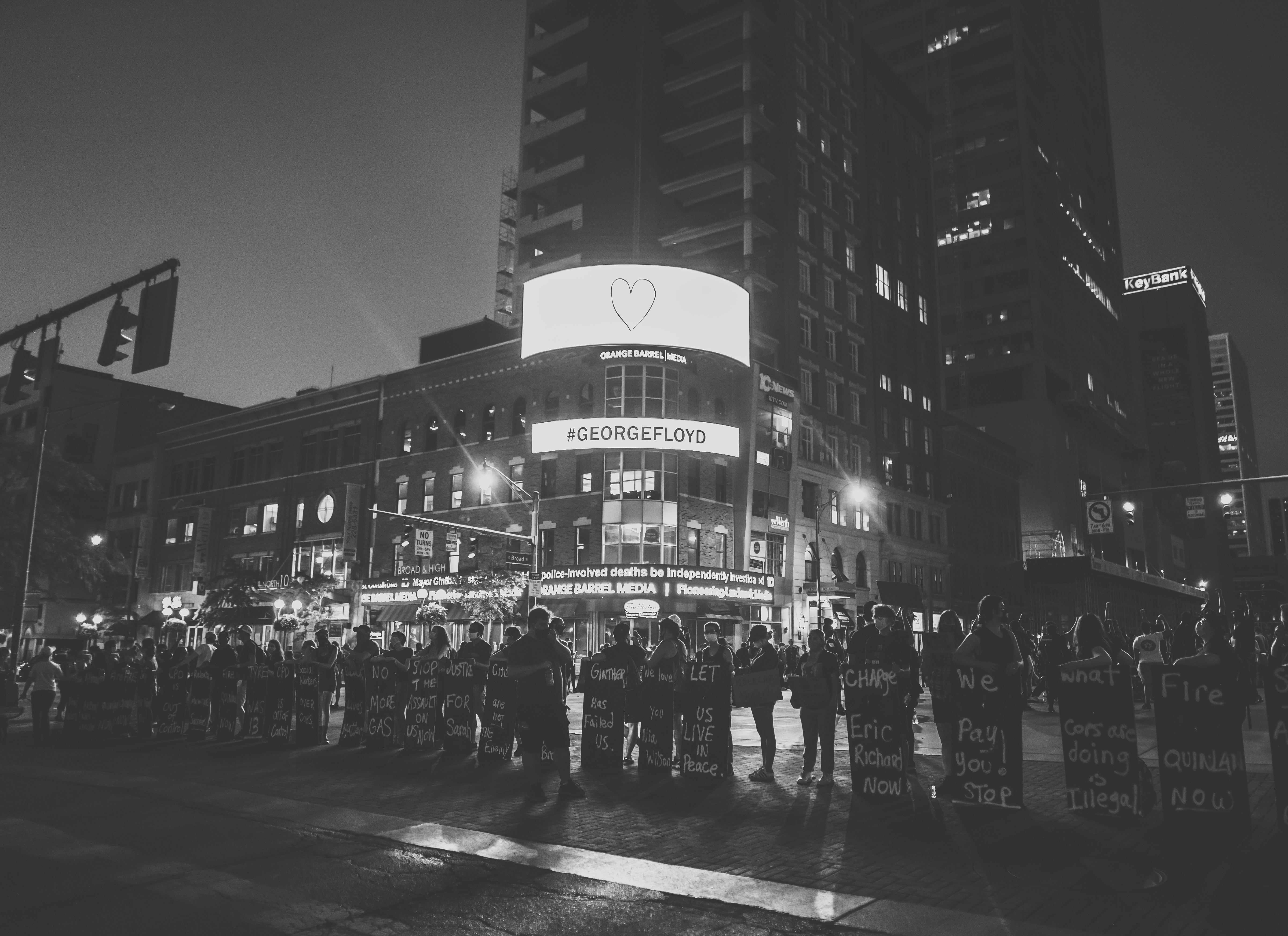 A black and white photograph. In a line across the bottom third of the image stand lines up BLM protesters that are holding chest-high signs in downtown Columbus at night. The background shows a building with a LED screen that reads "#GeorgeFloyd."