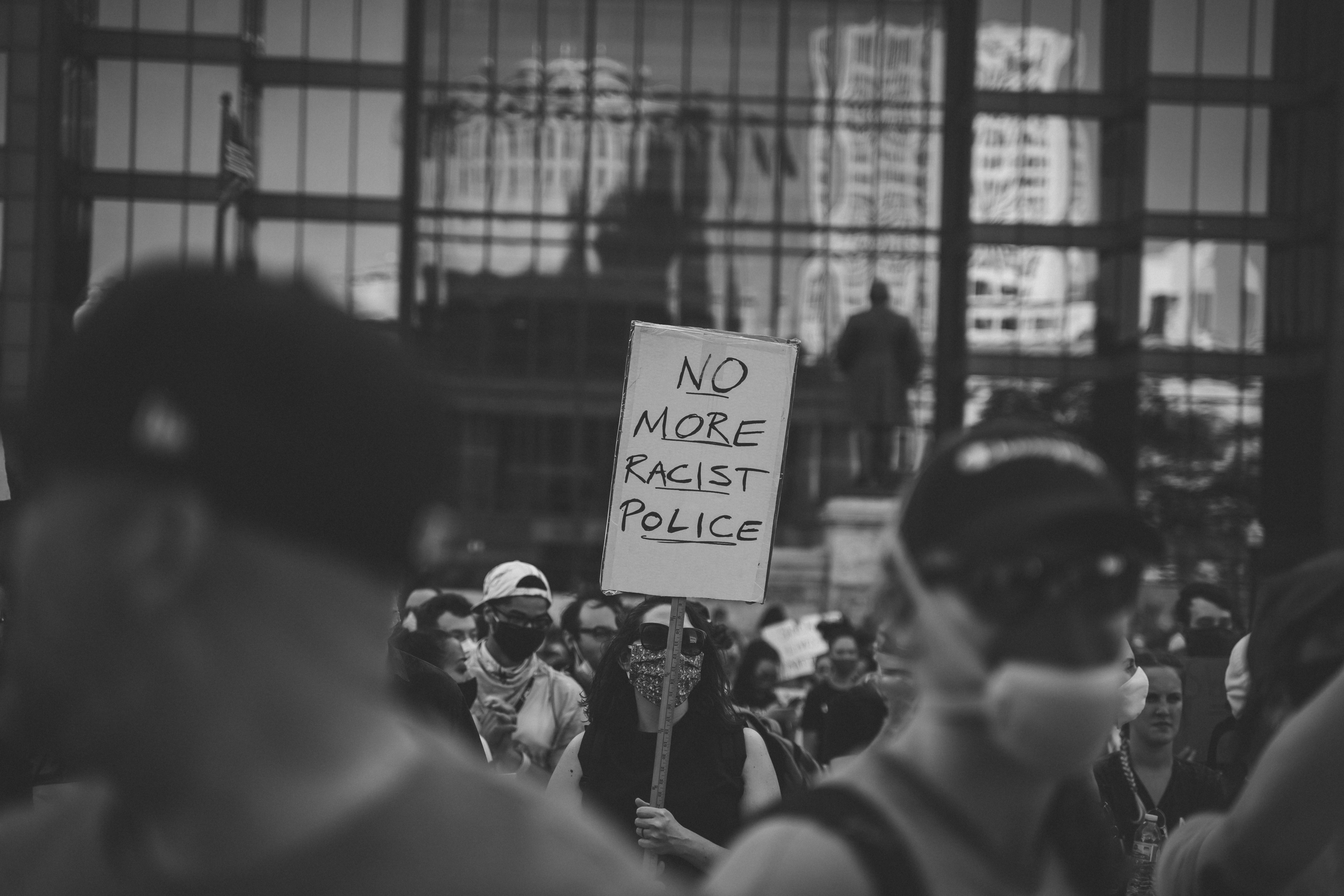 A black and white photo. The image focuses on the middle ground featuring a protester carrying a sign that says "no more racist police." The foreground shows more protestors out of focus. The background shows a building with windows that reflect images of many buildings in downtown Columbus.