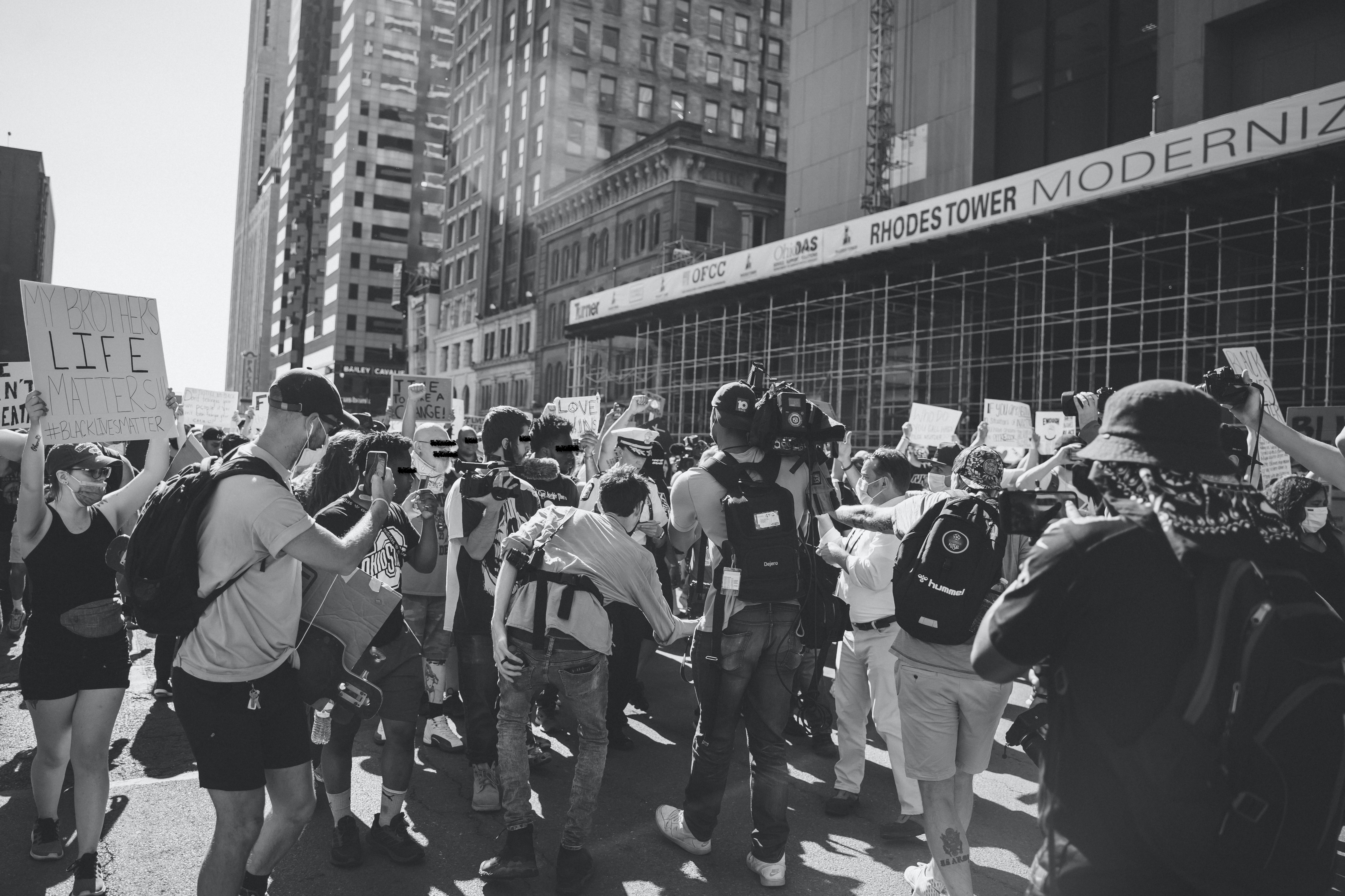 A black and white photograph. The bottom middle half of the image has a group of protestors with camera equipment and surrounding a police officer. BLM protesters with signs encircle the protestors with cameras. The top half of the image shows buildings in downtown Columbus.