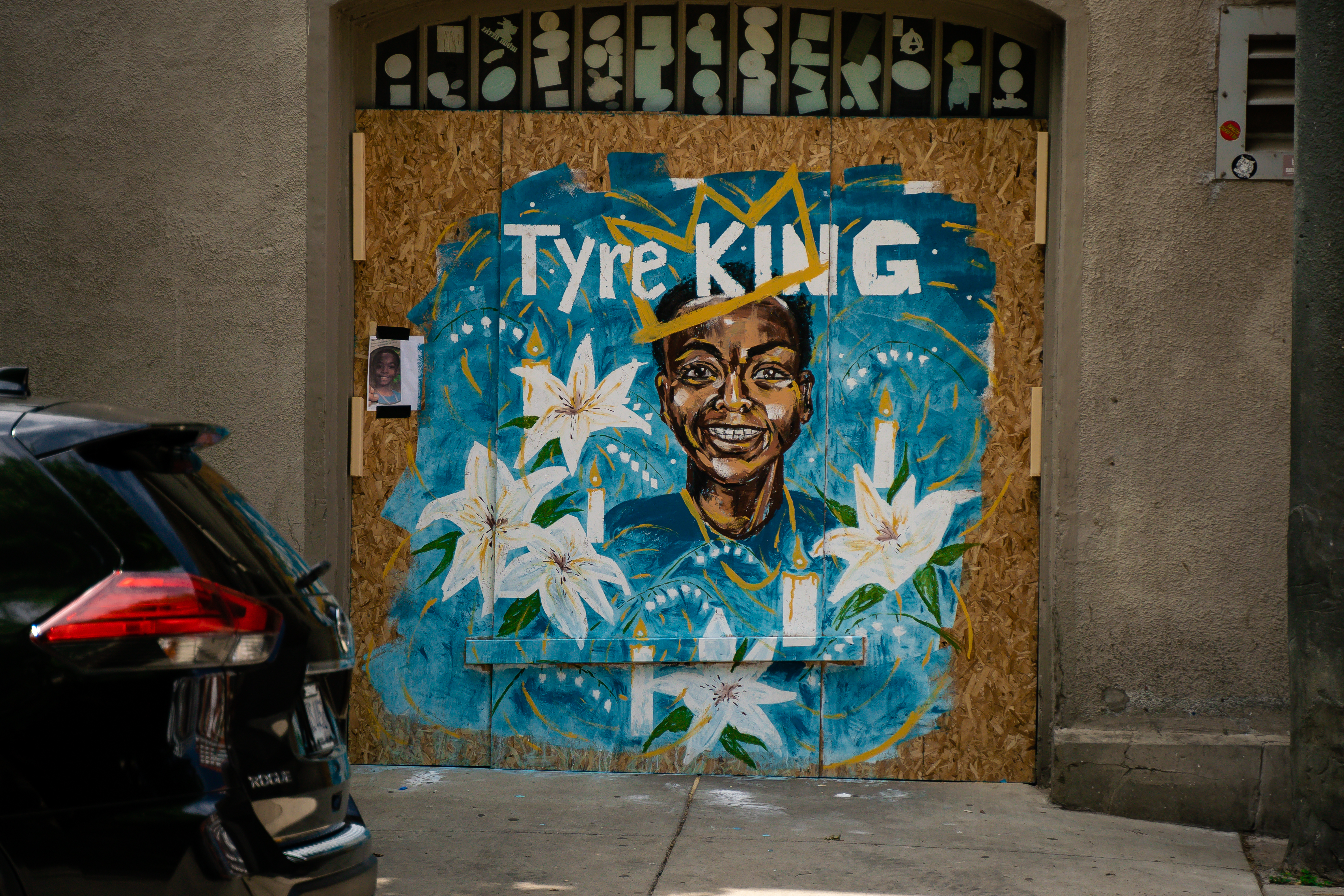 Street Art in Downtown Columbus on plywood covering windows on a building downtown Columbus. A bright blue background is painted on the plywood featuring the painted portrait of Tyre King. He is smiling and wears a golden crown, with the text "Tyre King" above the portrait. Next to the portrait are painted images of white flowers, lit white candles, and golden to honor Tyre King.