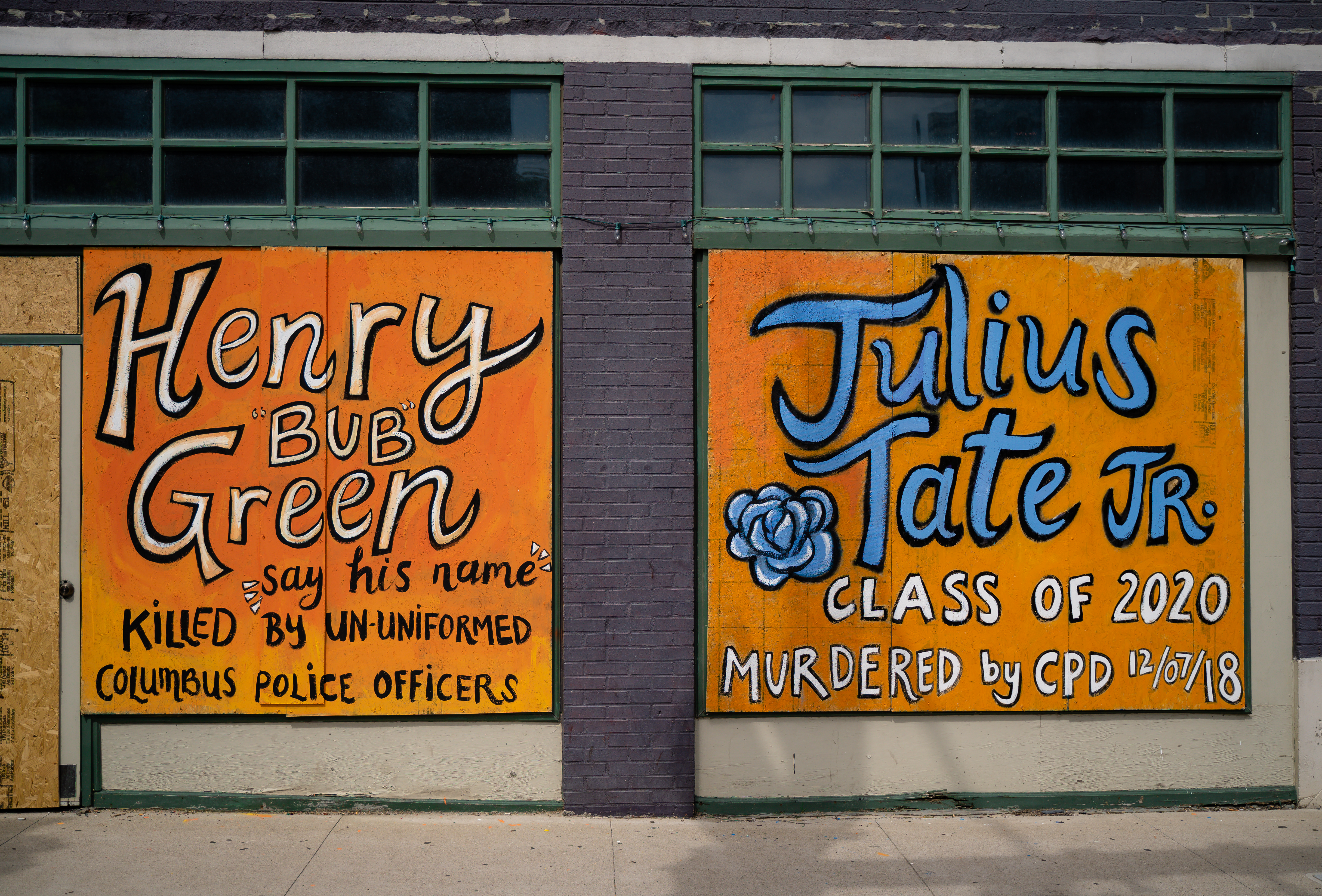 A color image of two square pieces of street art downtown Columbus on plywood over windows. The diptych both feature bright orange backgrounds. The left one reads in blacks and white colored text, “Henry ‘Bub’ Green,” “say his name,” and “killed by un-uniformed Columbus Police Officers. The right one reads in black, bright blue, and white text, “Julius Tate Jr.,” “Class of 2020,” “murdered by CPD 12/07/18.” Next to the word “Tate” is a black, blue, and white painted flower.