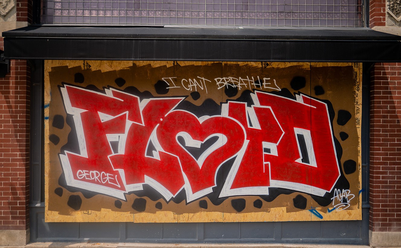 A color photo taken from head-on. The photo shows street art on wooden boards covering windows on a brick building downtown. The biggest word is FLOYD, with the "O" shaped as a heart is red, with white and black drop shadows. Three smaller white text blurbs appear across the red text and reads: George; I can't breathe; ACAB. "FLOYD" is on a brown background with black spots. 