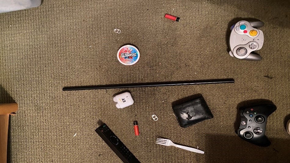 playstation controller, wallet, plastic fork, icebreaker candy, lighter, battery, metal rod laid out on brown carpet
