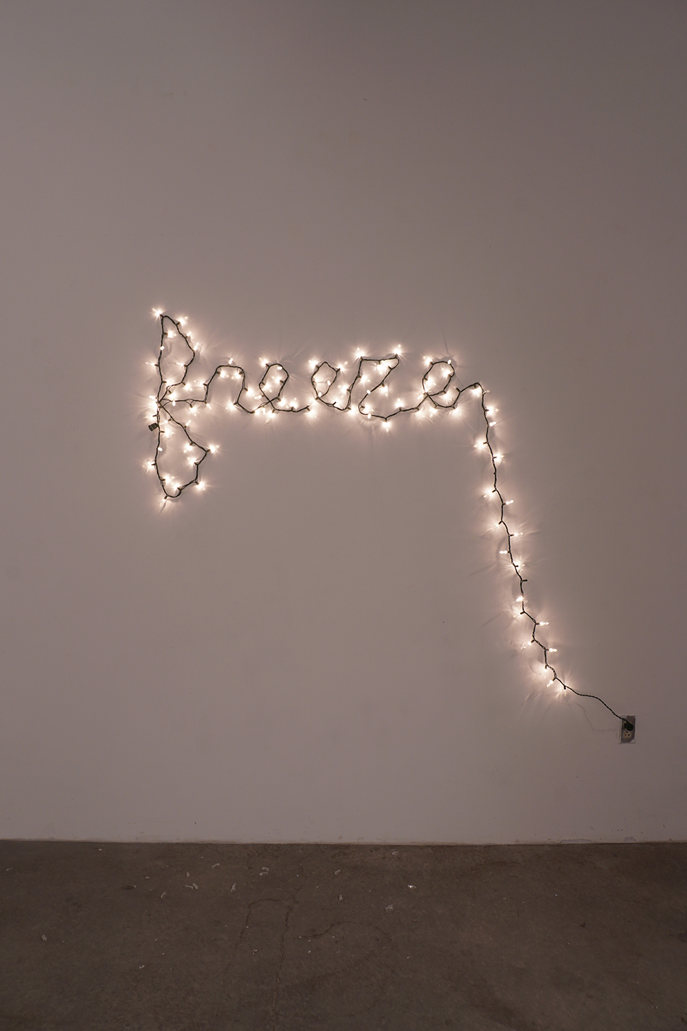 A photograph of string lights lit up, shining in a warm white color, and hung on a wall to spell the word 'freeze' in lowercase cursive. The end of the last 'e' curves downward leading down to an electrical socket.