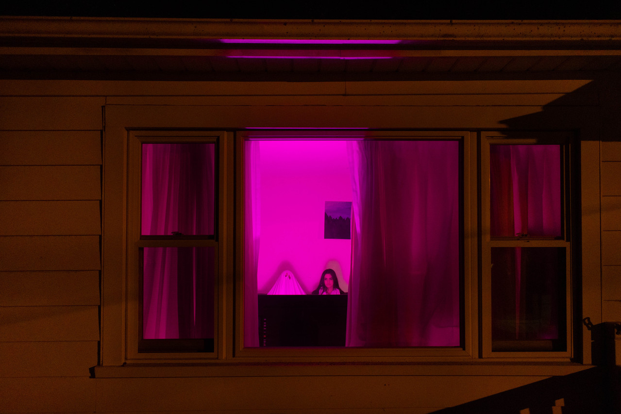 photograph of two people, one dressed as a ghost, sitting in a living room while bathed in a neon violet light, the viewer is seeing this through the house's window