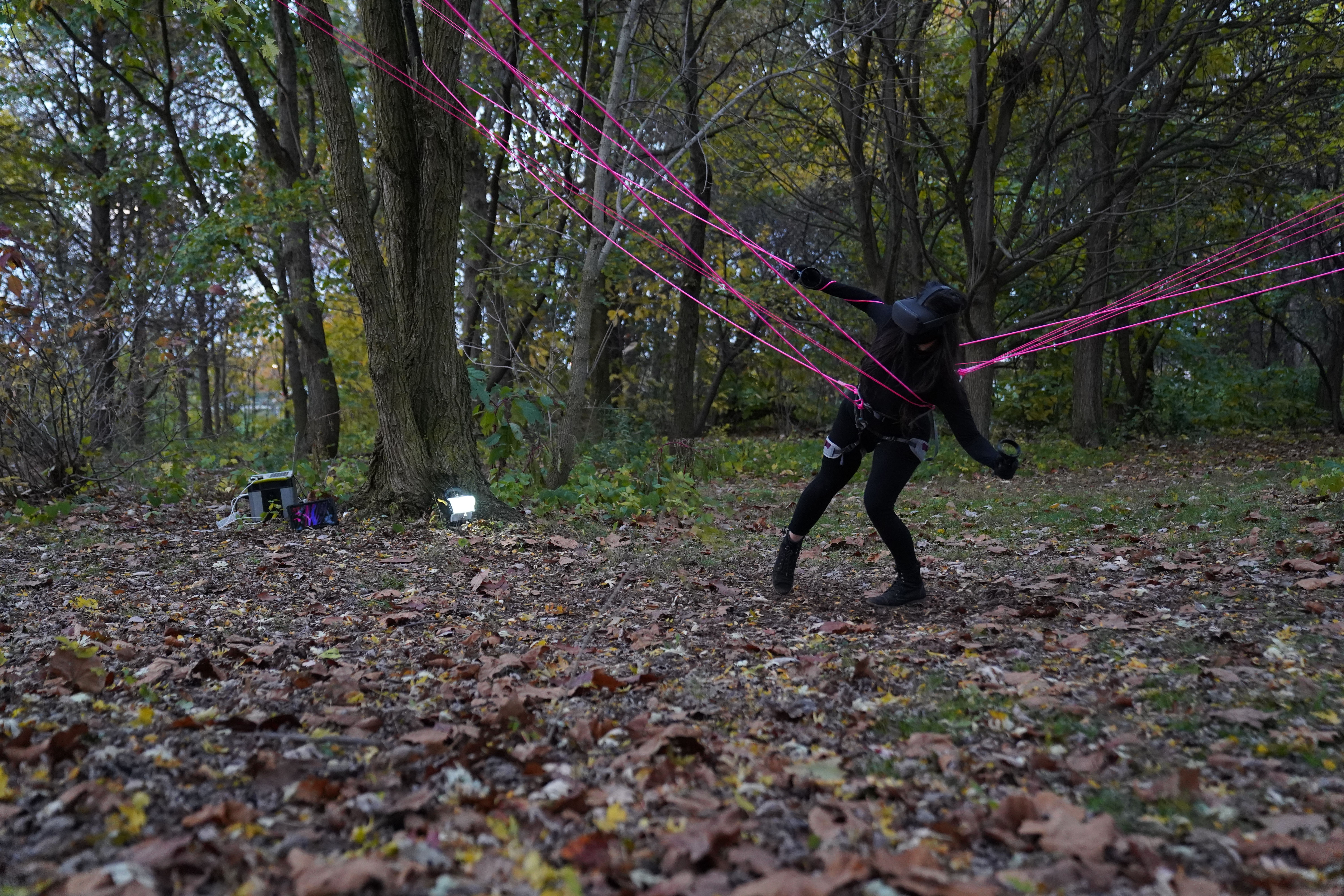 A figure wearing all black and a VR headset is caught in webs of neon pink string that is strung between trees in a wooded area. Far on the left, out of reach from the figure is digital equipment. and a light