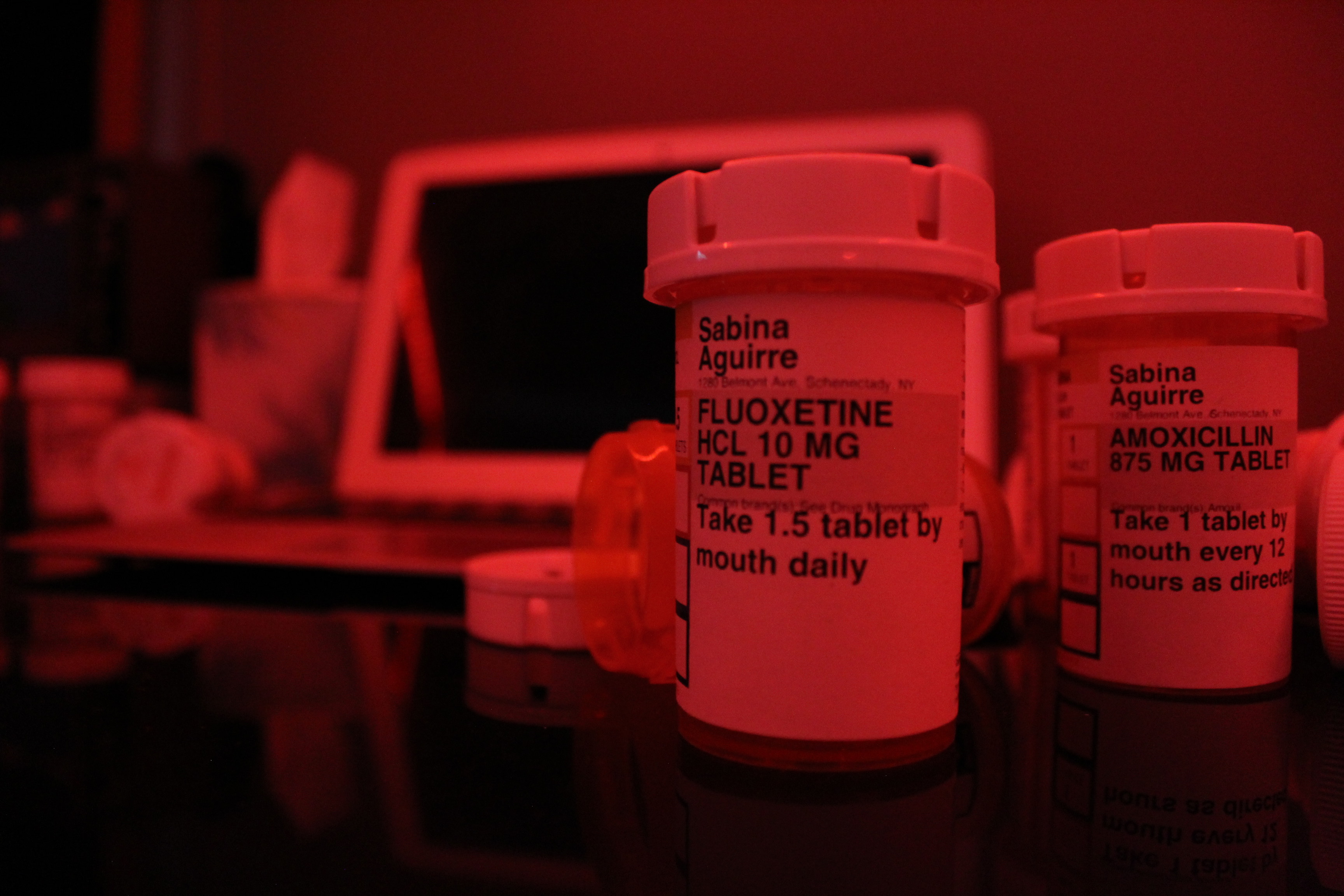 A red-tinted close-up photo of two pill bottles, labeled "Fluoxetine" and "Amoxicillin," prescribed to Sabina Aguirre