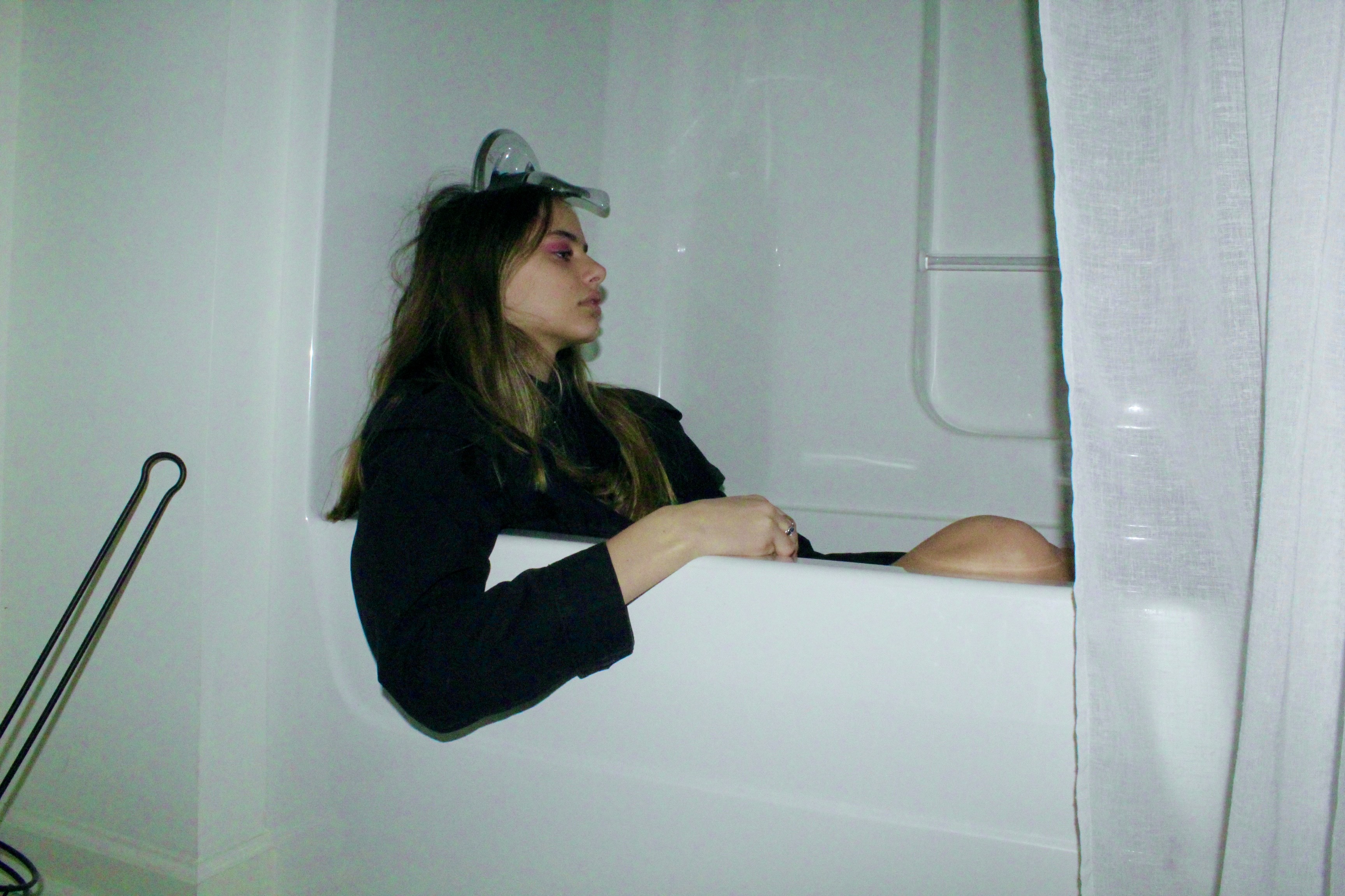 A person with messy hair and pink eyeshadow sits fully-clothed in a bathtub