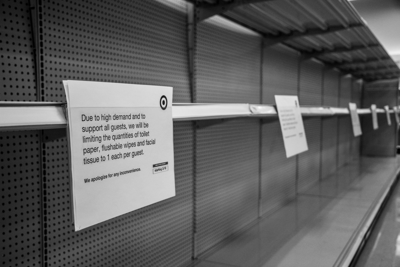 A black-and-white photo of empty store shelves with signs informing guests the amount of paper products they can buy are limited