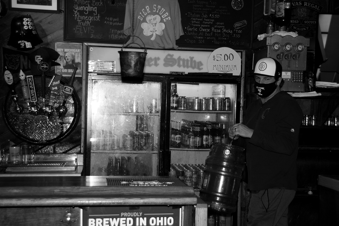 A black-and-white photo of a person wearing a mask and holding a keg behind the Bier Stube bar