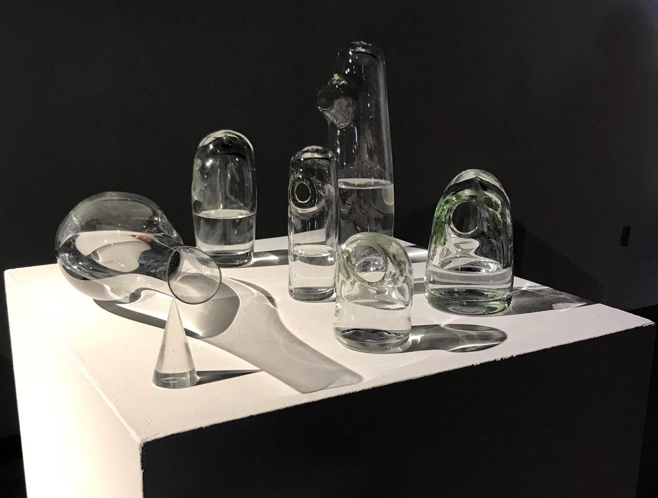 Seven pieces of glassware, most filled with water, sit on a platform in front of a black background