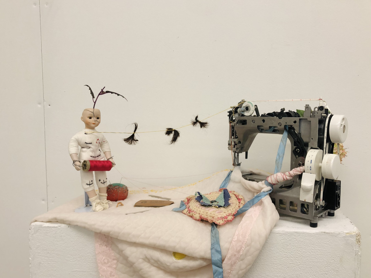 A baby doll covered in black stitches holds a spool of red thread while standing on a light pink blanket. Sewing supplies are strewn across the blanket. To the right is a sewing machine with no outer casing.
