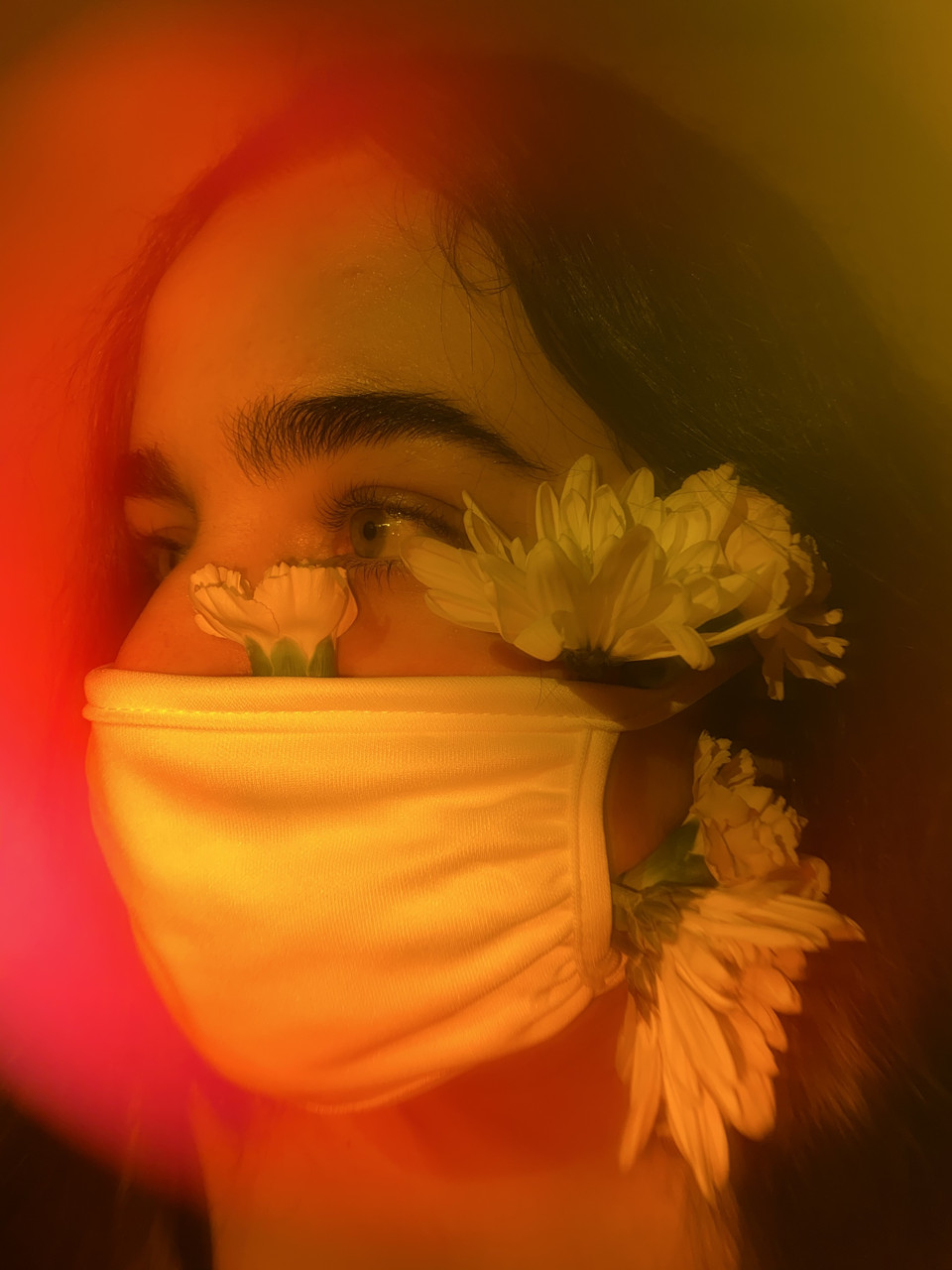 An orange-tinted close-up photograph of a person wearing a mask with yellow flowers tucked inside