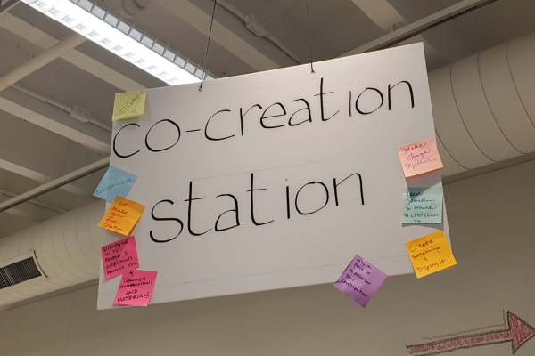 A sign hanging from the ceiling of the gallery stating "Co-creation Station." Colorful sticky notes adorn the edges of the sign.