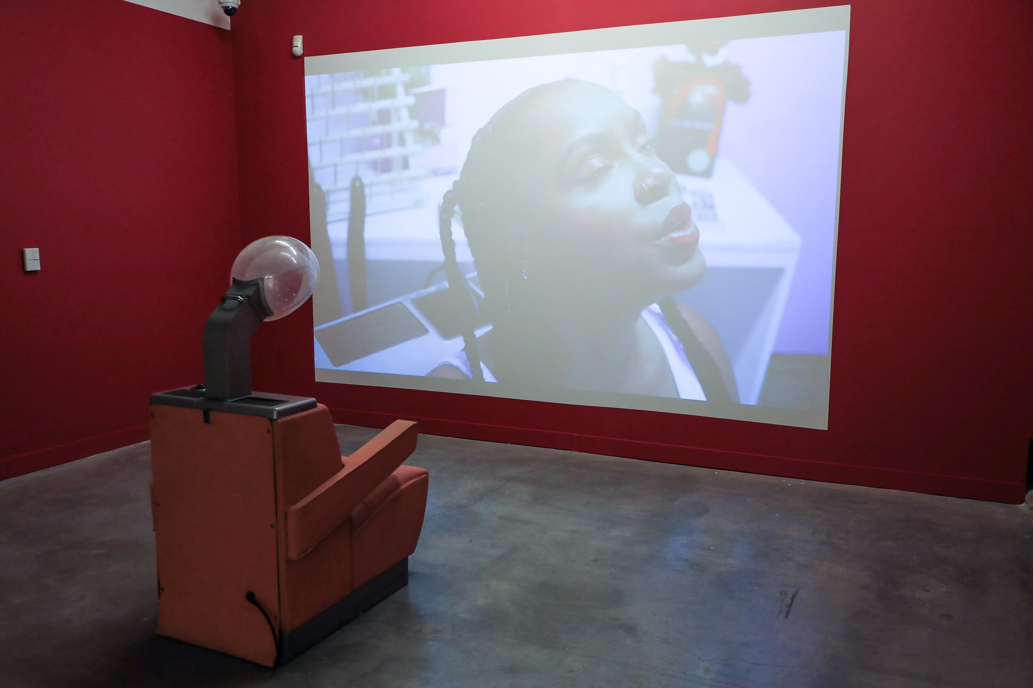 Video projection in red painted room. A recliner with a hair dryer attachment watches the video. 