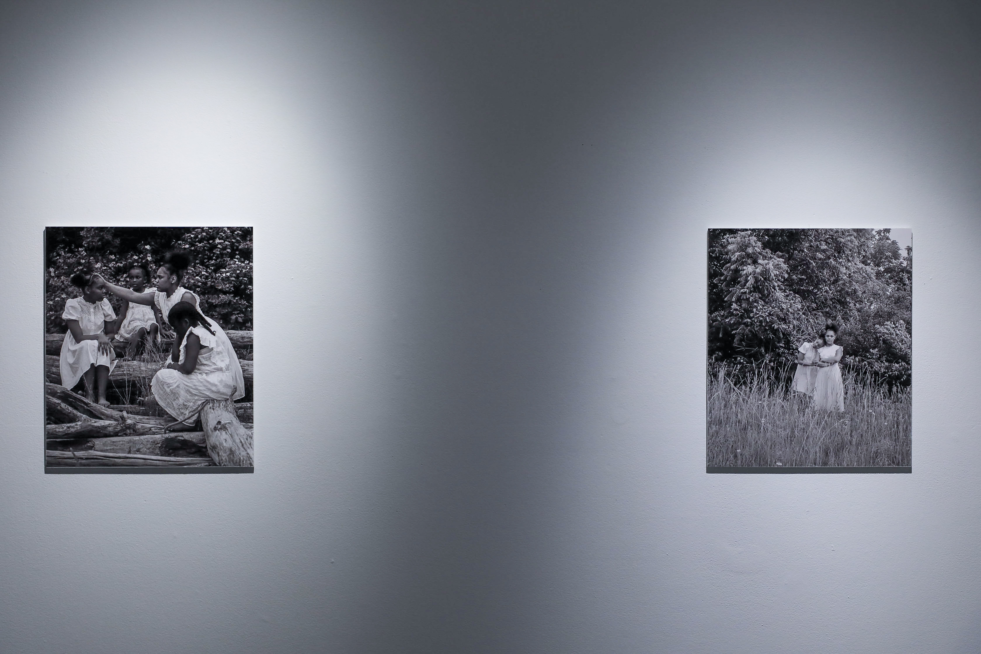 There are two black and white photos: the one on the left shows four black girls dressed in white sitting on logs with one black girl reaching out to another. The one on the right shows two black girls dressed in white holding each other in a field with a tree in the back. 