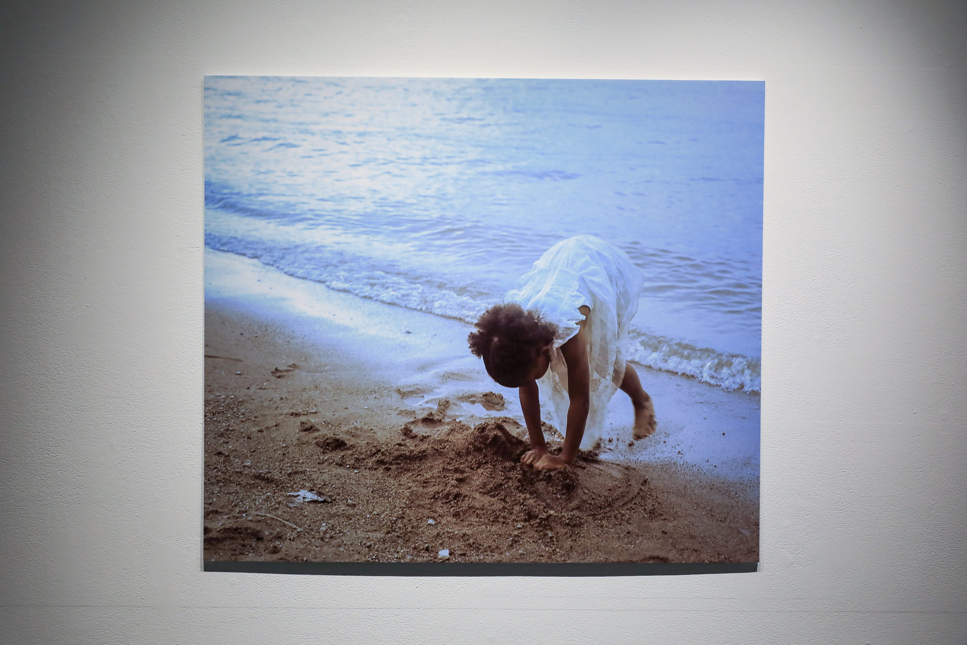 A color photo of a black girl dressed in white playing with sand on a beach.