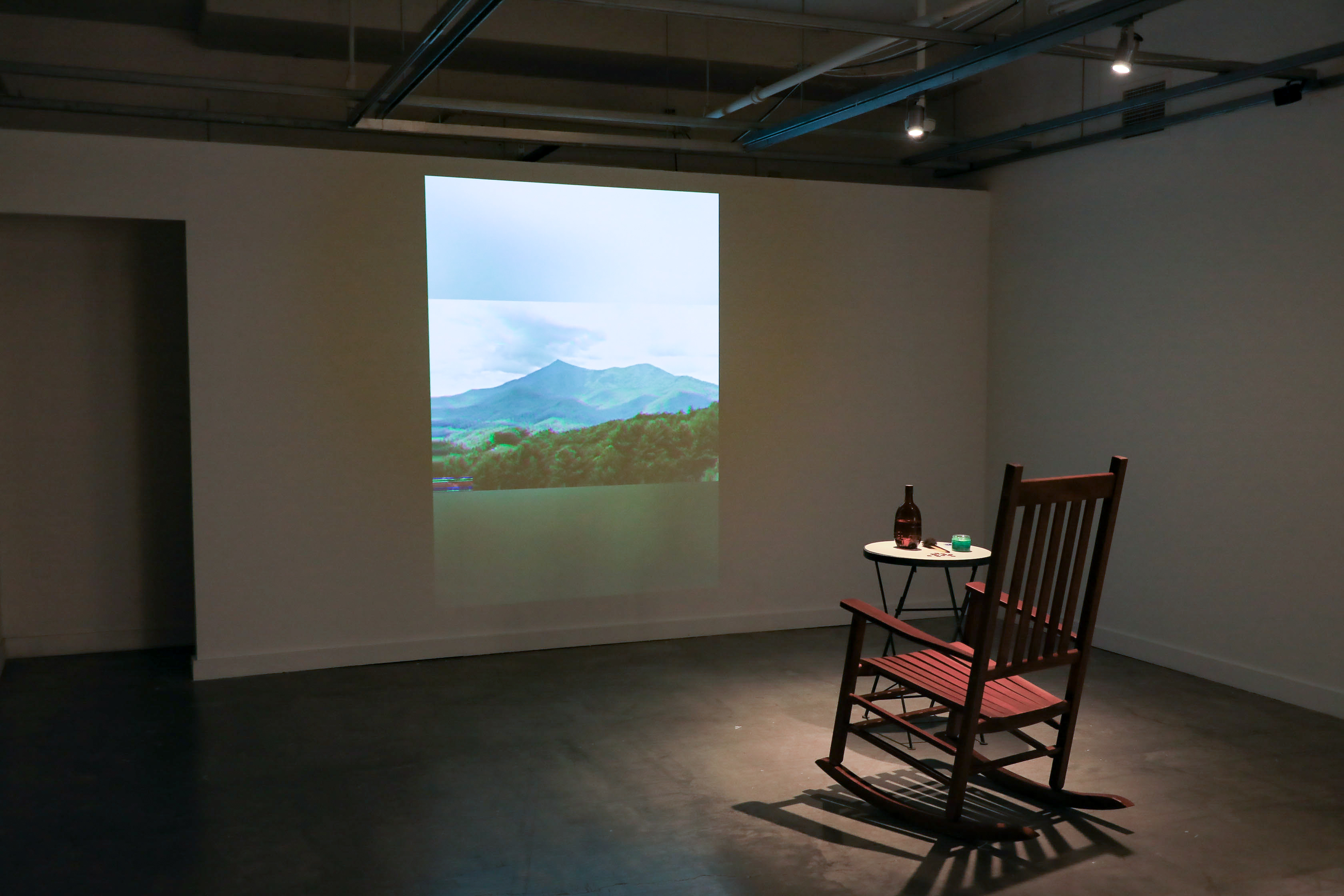 A rocking chair in front of a projected video of nature. To the right of the rocking chair is a table with a bottle and assorted items. 