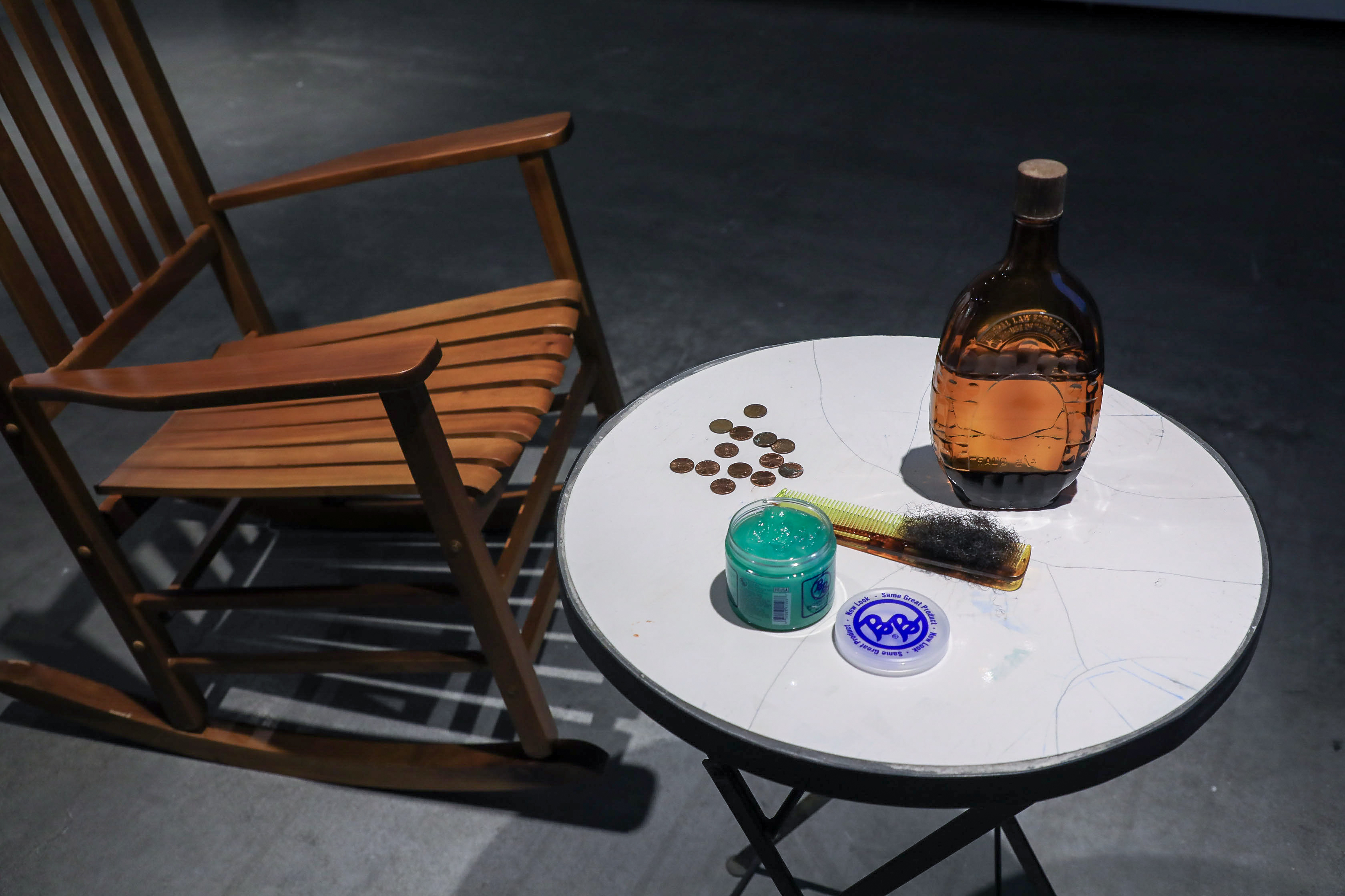 A close-up of the rocking chair and table: the table has a bottle with brown liquid, a brush, pennies, and hair gel. 