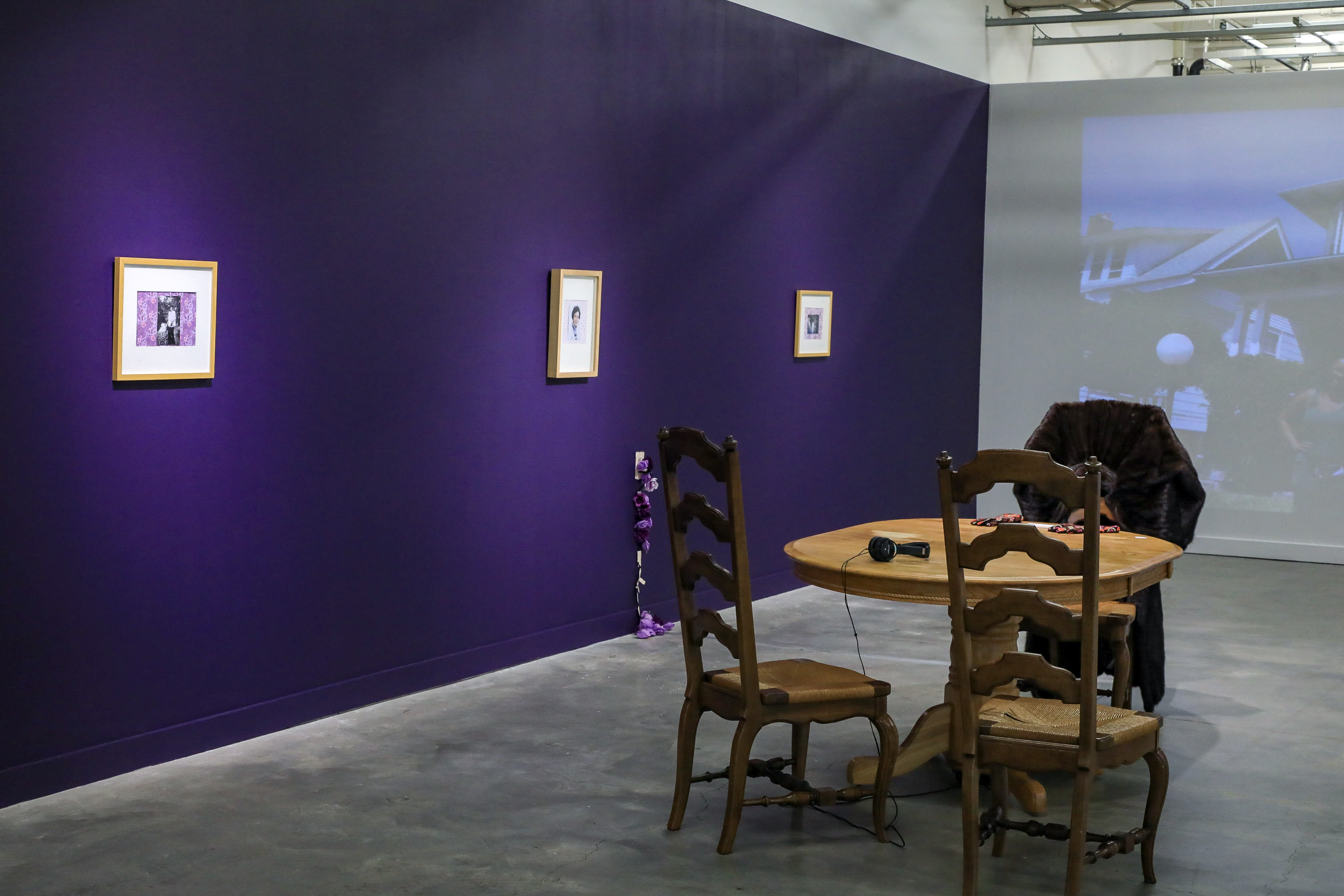 Three framed photos on a purple wall-on the adjacent wall there is a projected video. In the middle of the area there is a table with three chairs; a mink coat is draped over main chair.