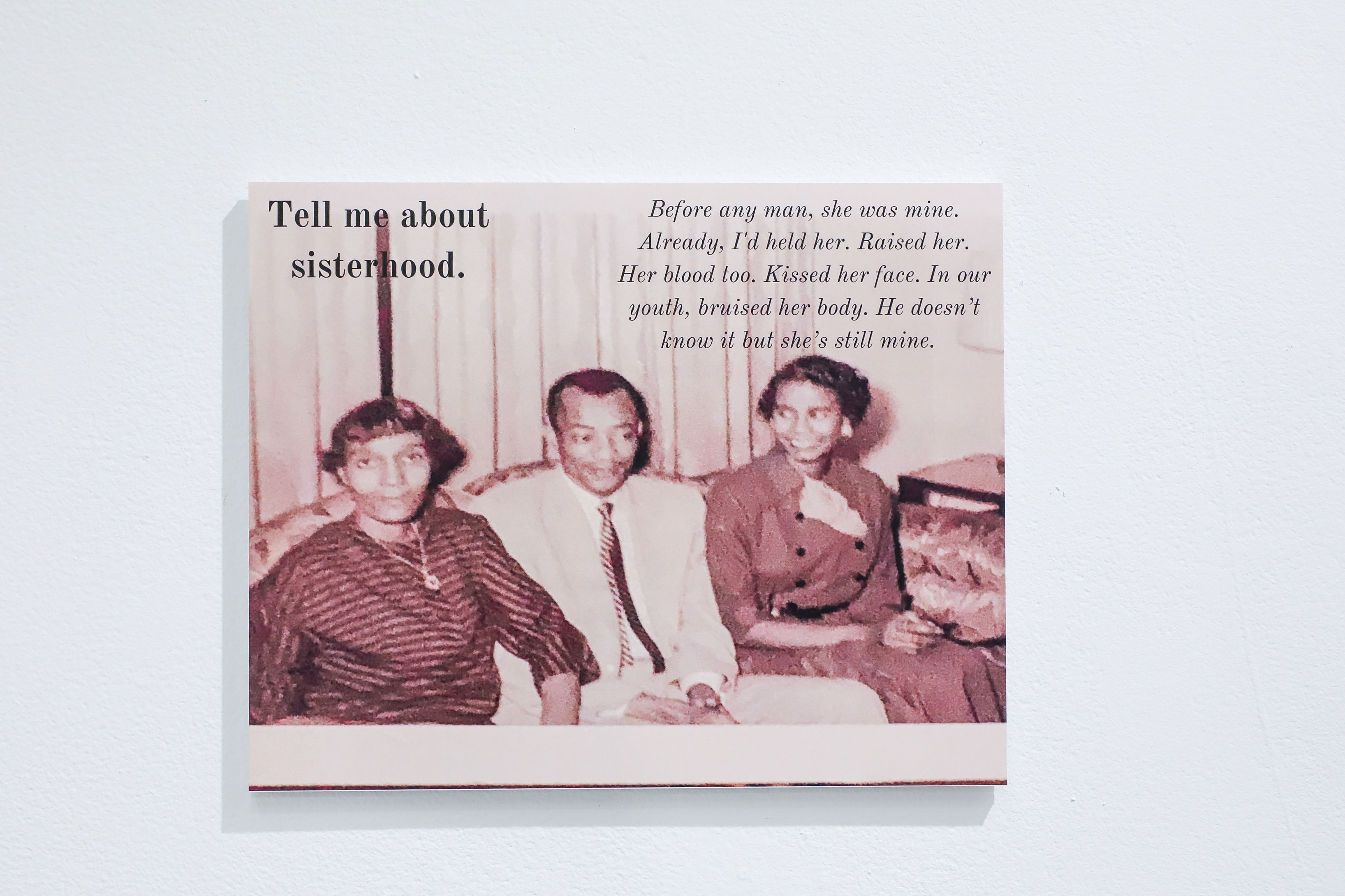 A photo of two black women and a black man in between them sitting together. The text says “Tell me about sisterhood. Before any man, she was mine. Already, I’d held her. Raised her. Her blood too. Kissed her face. In our youth, bruised her body. He doesn’t know it but she’s still mine.”