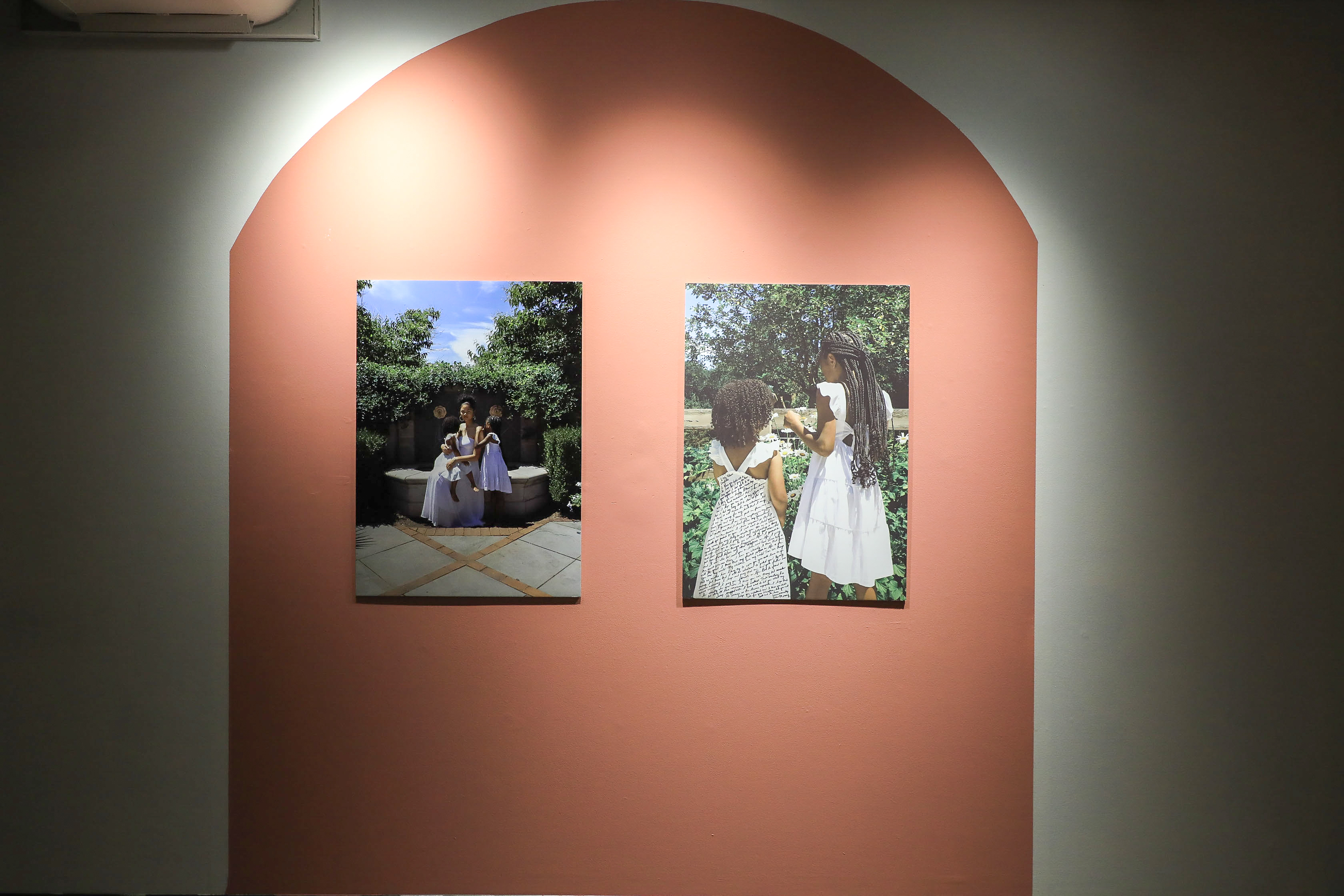 There are two color photos on top of a red-orange painted shape: the one of the left shows Iyana dressed in white sitting on a garden fountain with two little black girls next to her. The one on the right shows the little girls dressed in white standing in front of flowers with writing on the left girl’s dress. 