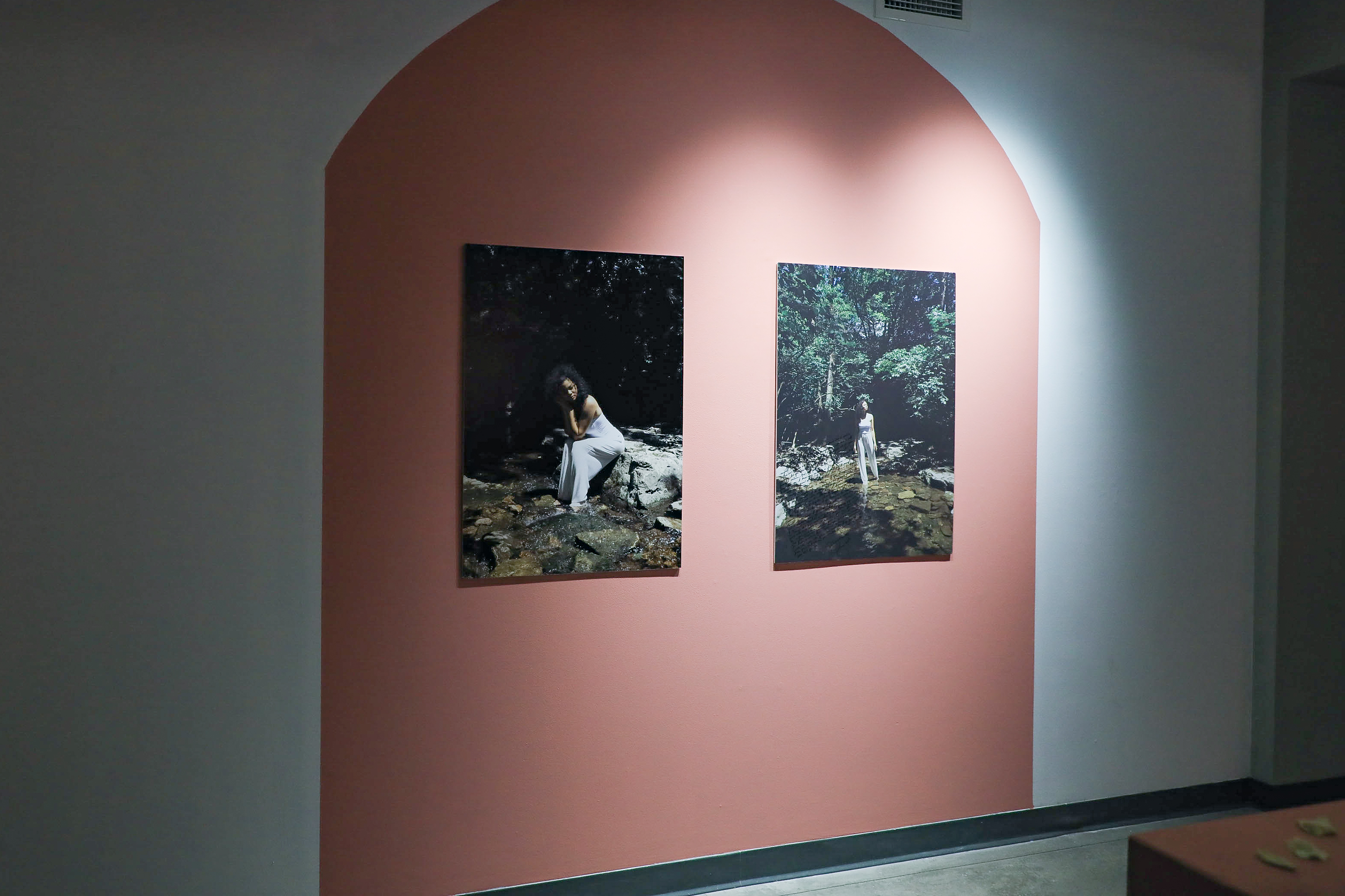 Two color photos on top of a red-orange painted shape: the one on the left show Iyana dressed in white sitting on a rock in a creek. The one on the right show Iyana standing in the creek with the woods in the background.