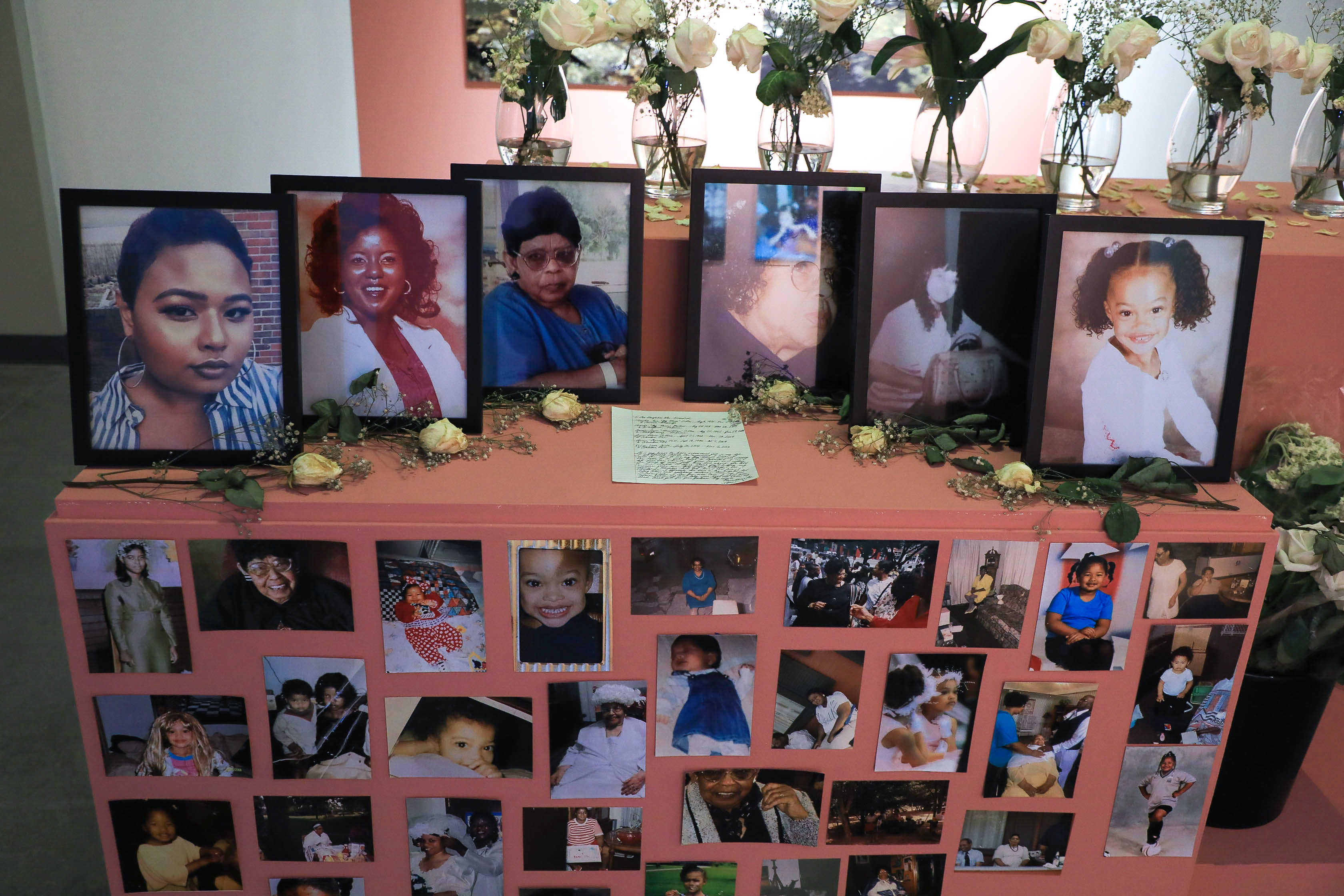 A red-orange painted box with dozens of photos decorating the box and six framed photos and flowers on top forming an altar. There are flowers on a ledge behind the framed photos and a letter in the middle. 