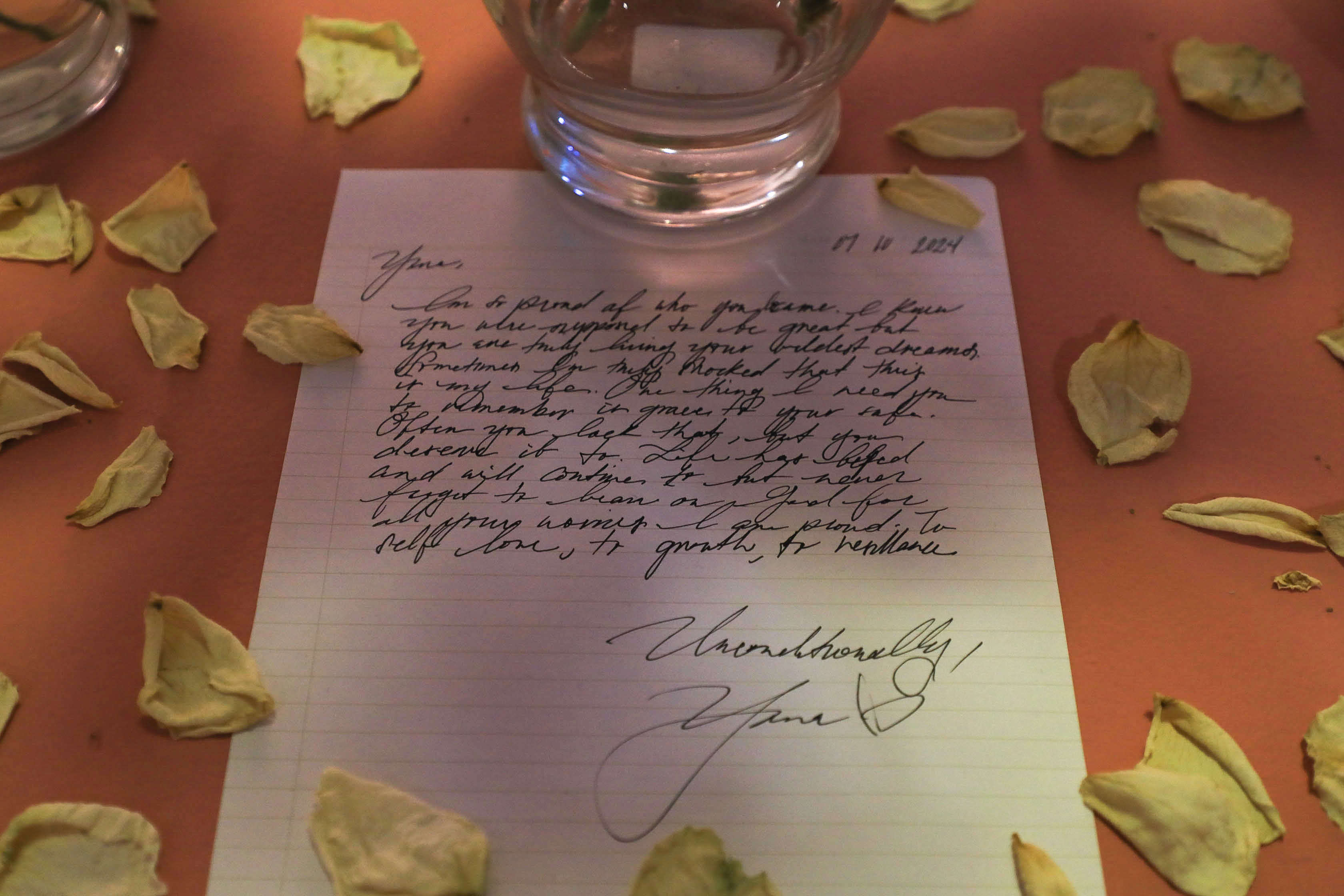 A letter with petals and vases surrounding it on a red-orange box.