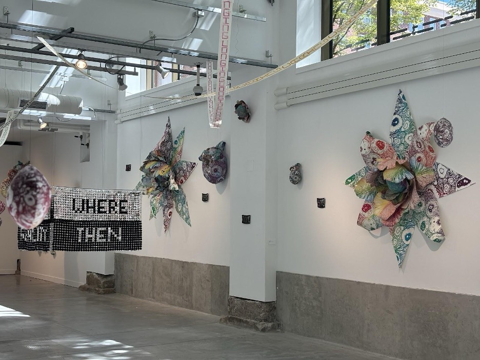 Alex McClay and Stephanie Berrie’s works are displayed attached to the wall. The two sculptures are vibrantly rainbow and form a 3-D flower shape. A metallic artwork suspends from the air, reading “WHERE, THEN” in the middle of the gallery