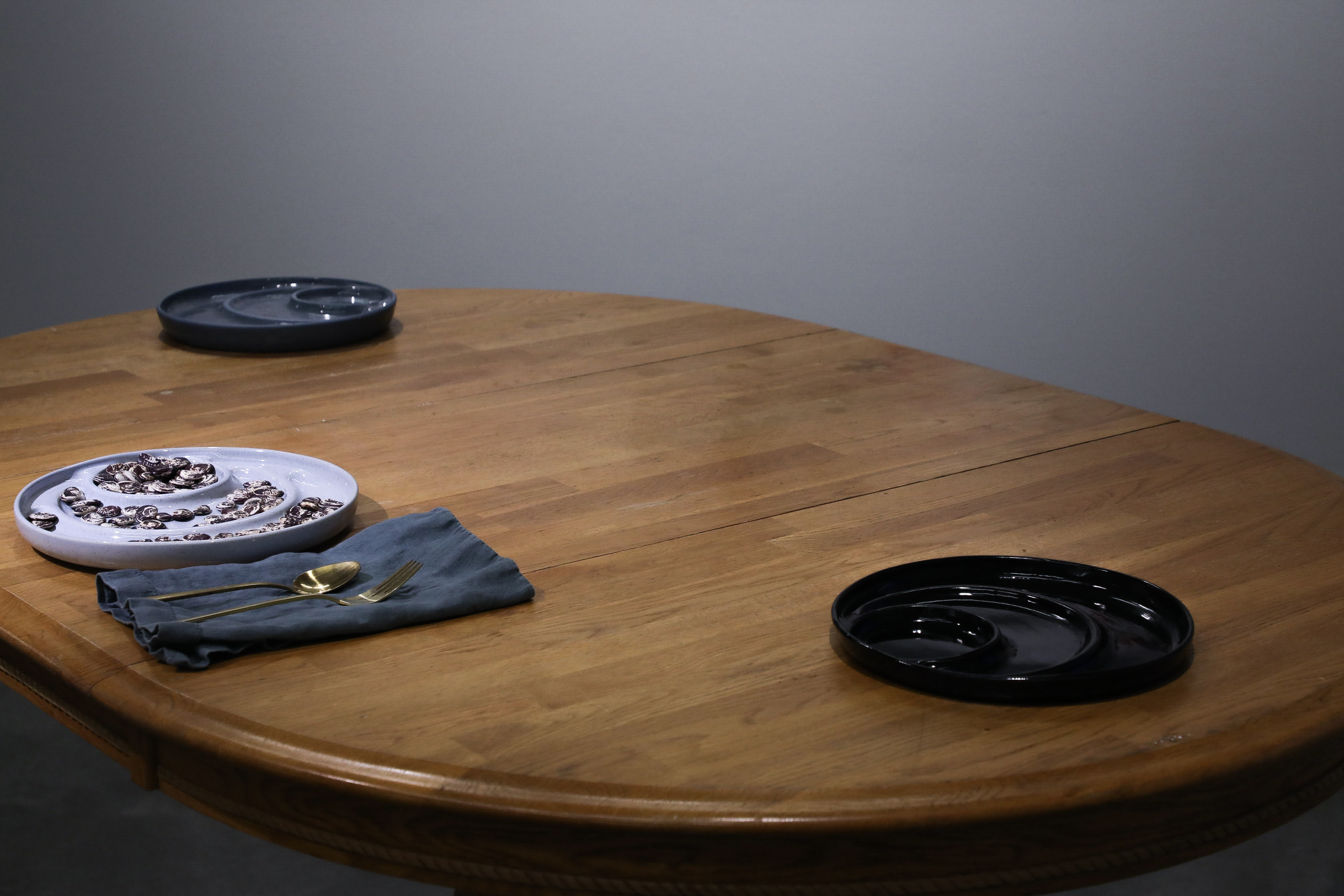 A wooden dining table that has three plates on it. One Plate is white,  the second is blue, and the last is black. The white plate has dried beans on it, and a set of utensils is next to it.