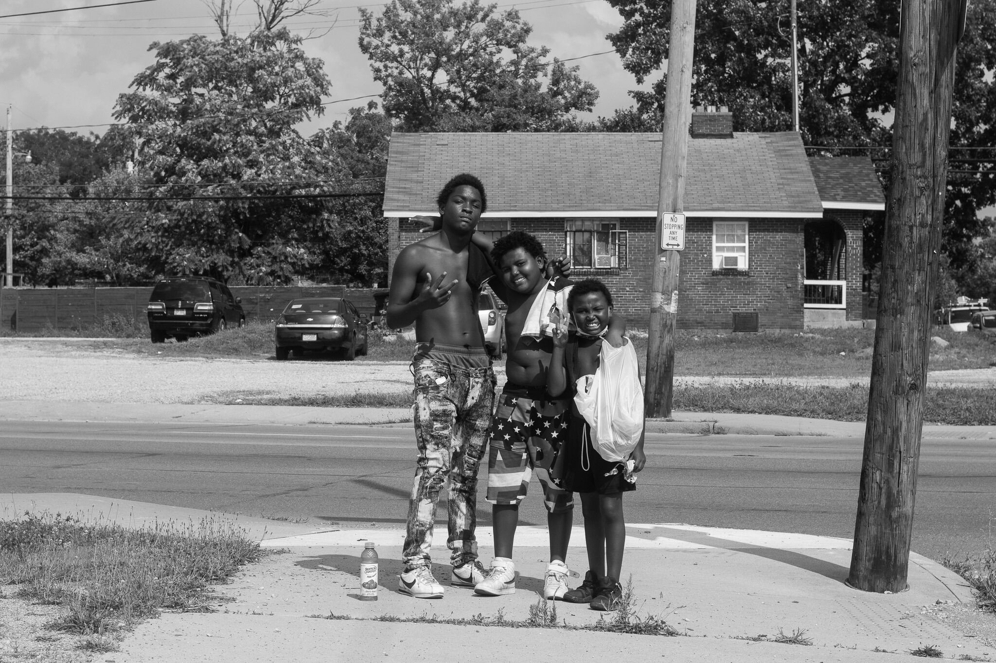 Three boys of color standing in order from tallest to shortest, all with shirts off and holding shopping bags 