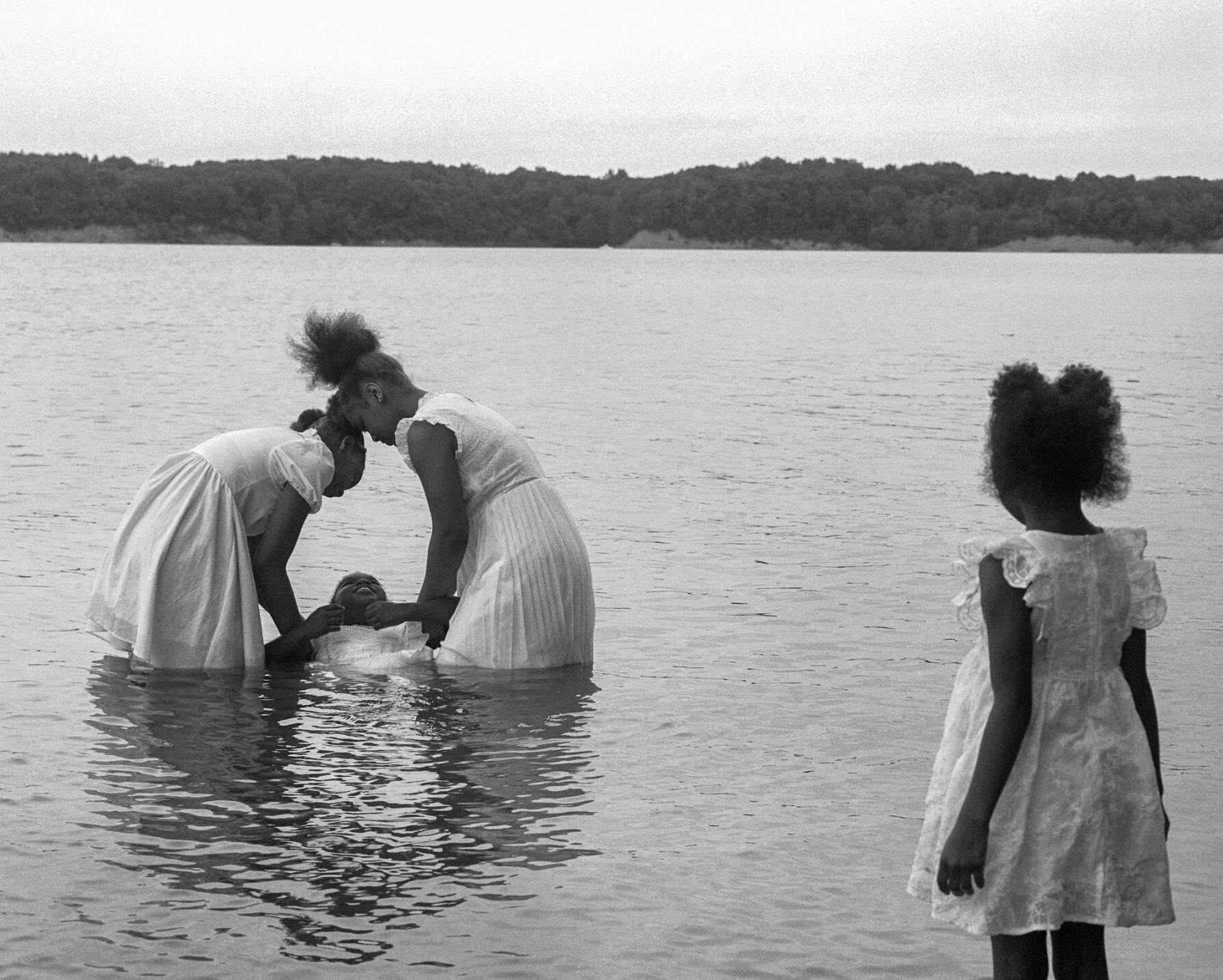 Four girls of color wearing white dresses standing in water. Two girls peacefully lower a third girl into the water as the fourth watches in the foreground.