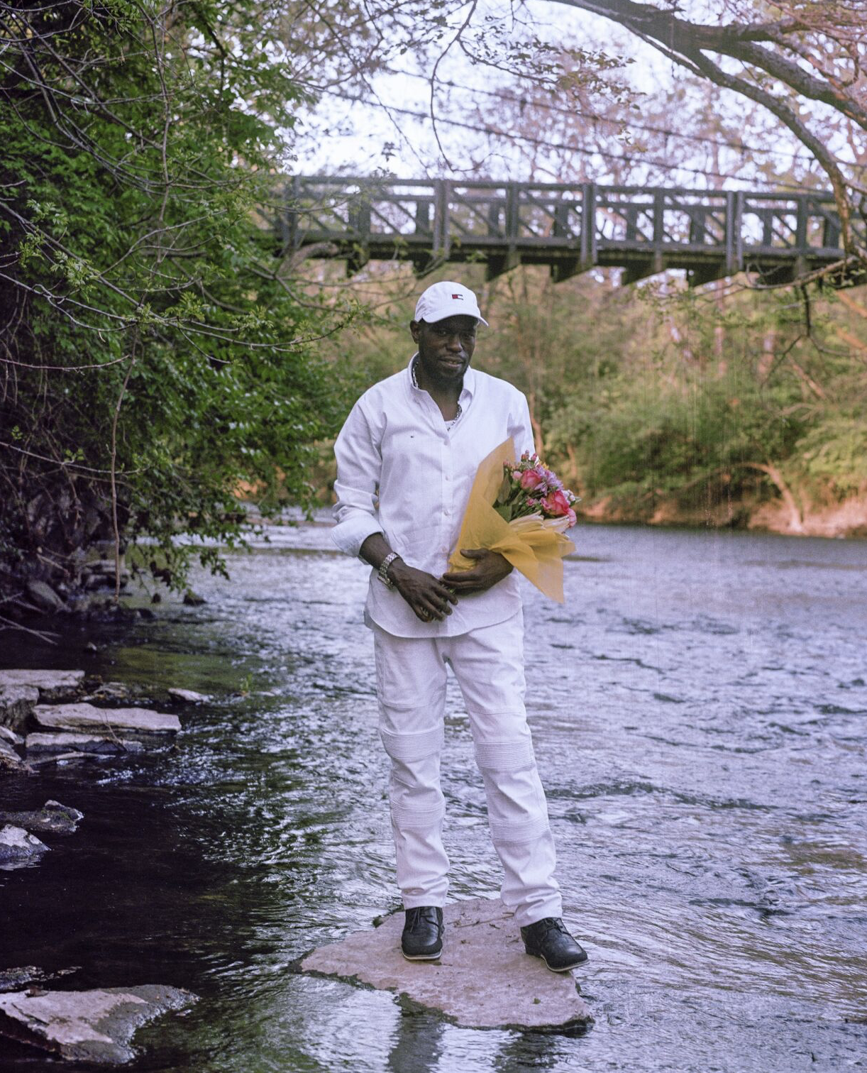 A man of color in a white outfit standing on a rock in a river in front of a brdige holding a bouquet of flowers