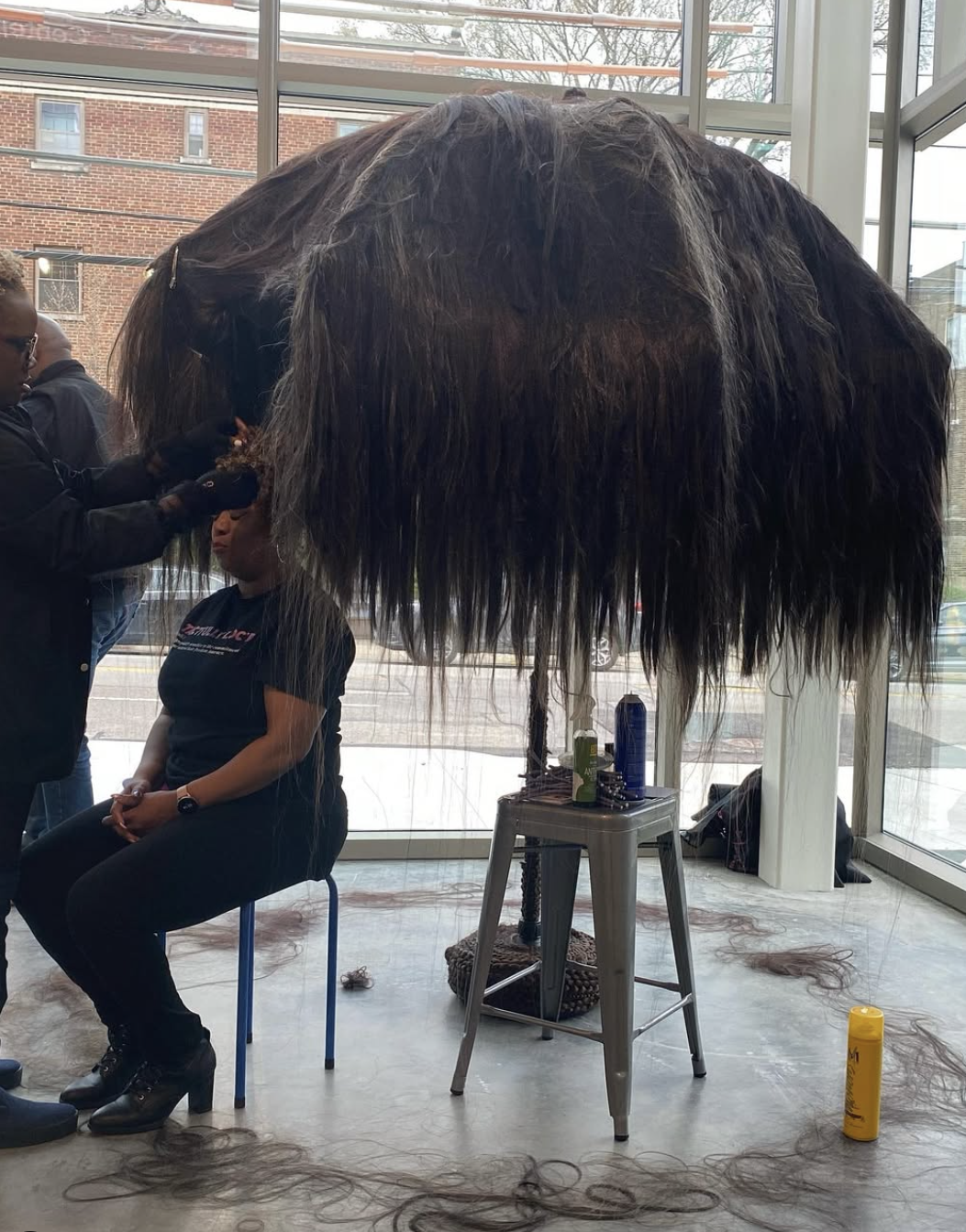 Photograph of a woman of color getting her hair done by another woman of color under a large, tree-like hut made from synthetic hair. 