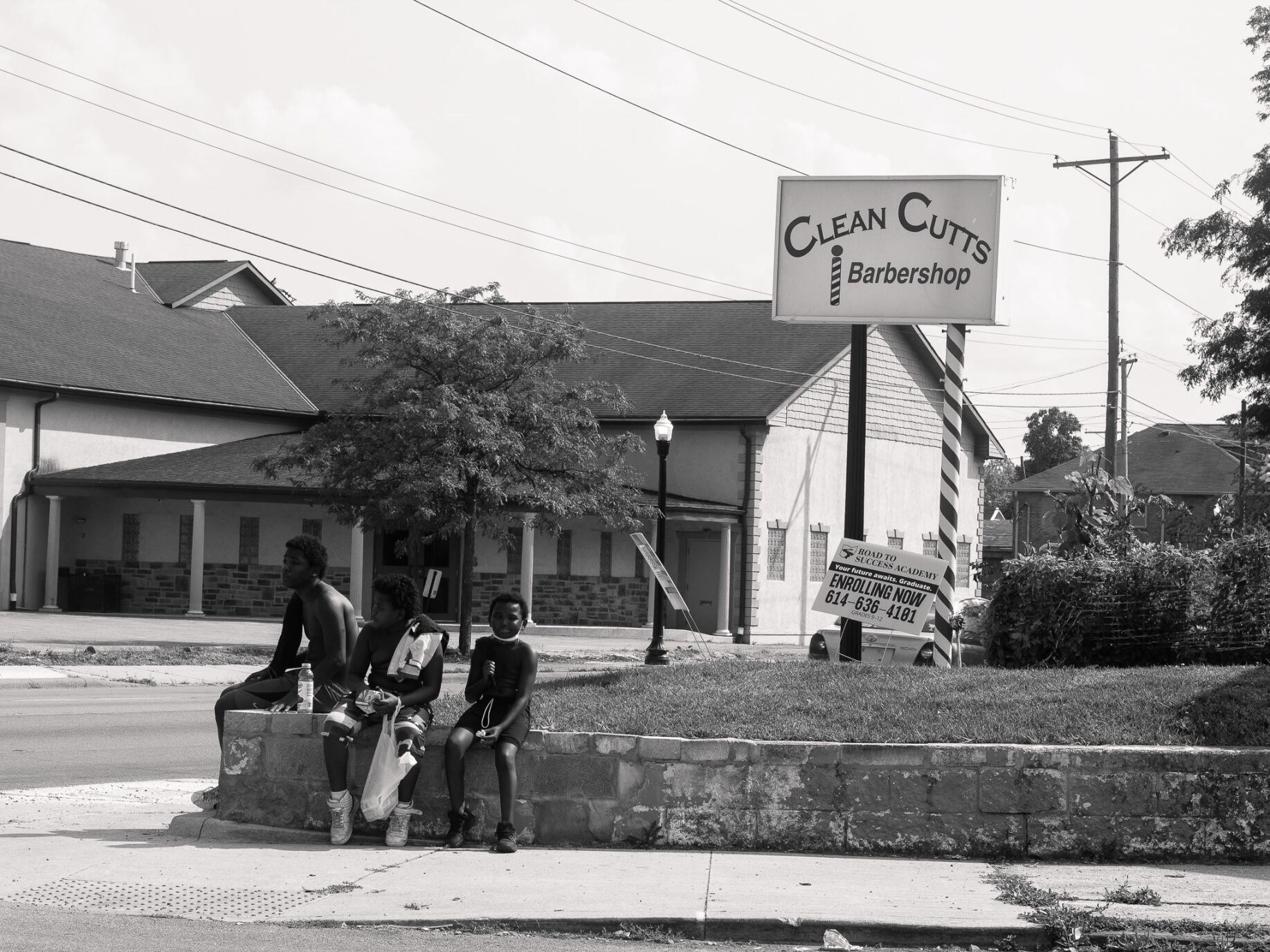 Three boys of color sitting on a wall on the side walk holding shopping bags in front of a sign called "Clean Cuts Barbershop"