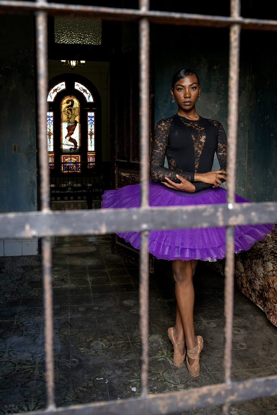 a woman of color in a purple tutu, black top, and ballerina shoes standing at point behind bars and in front of a stained glass window