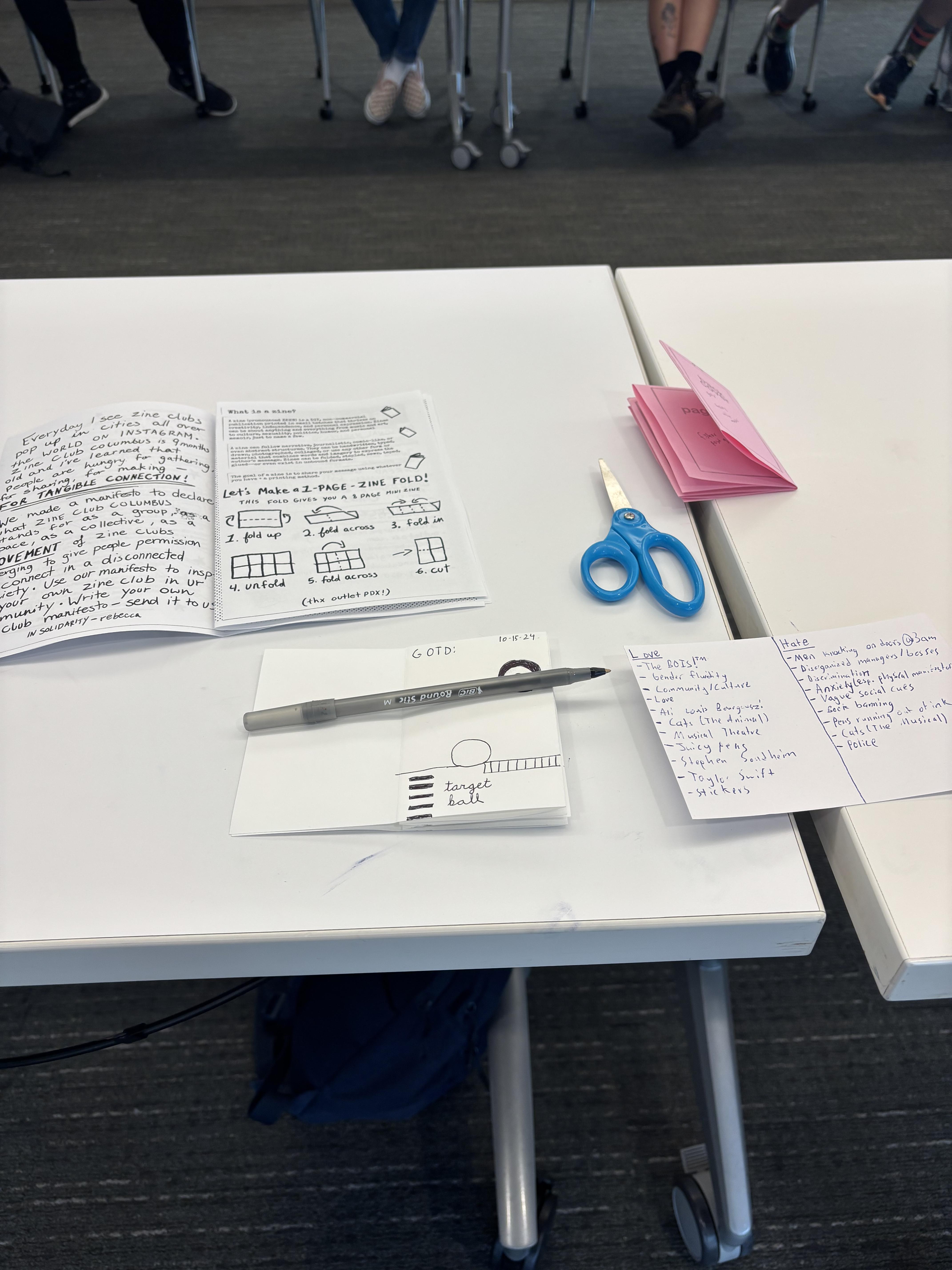 Instructions, a list on a note card, a one-page zine, a drawing, scissors, and a pen lay overlapping each other on a white table.