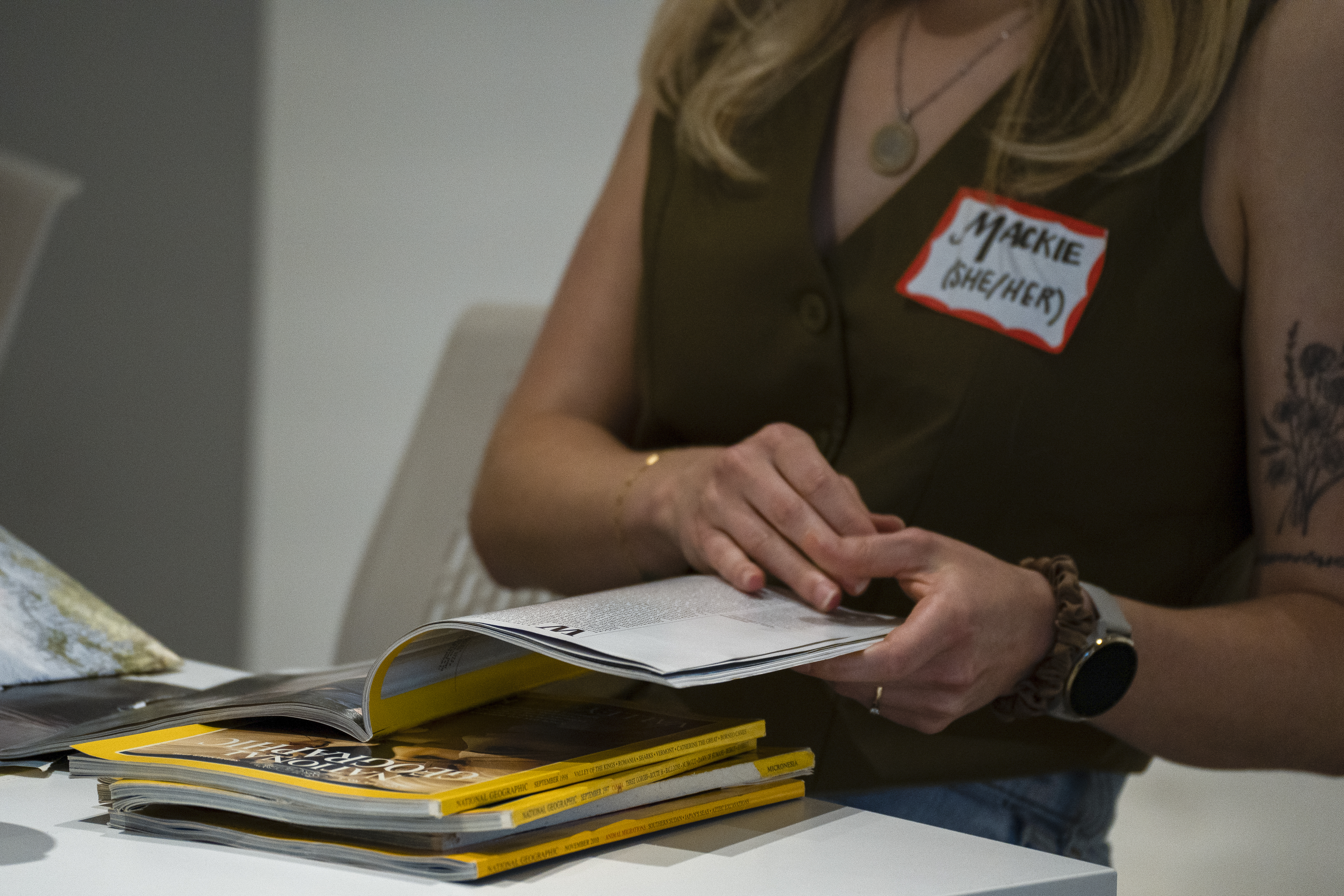 Woman wearing a name tag that says Mackie She/Her flips through a magazine.